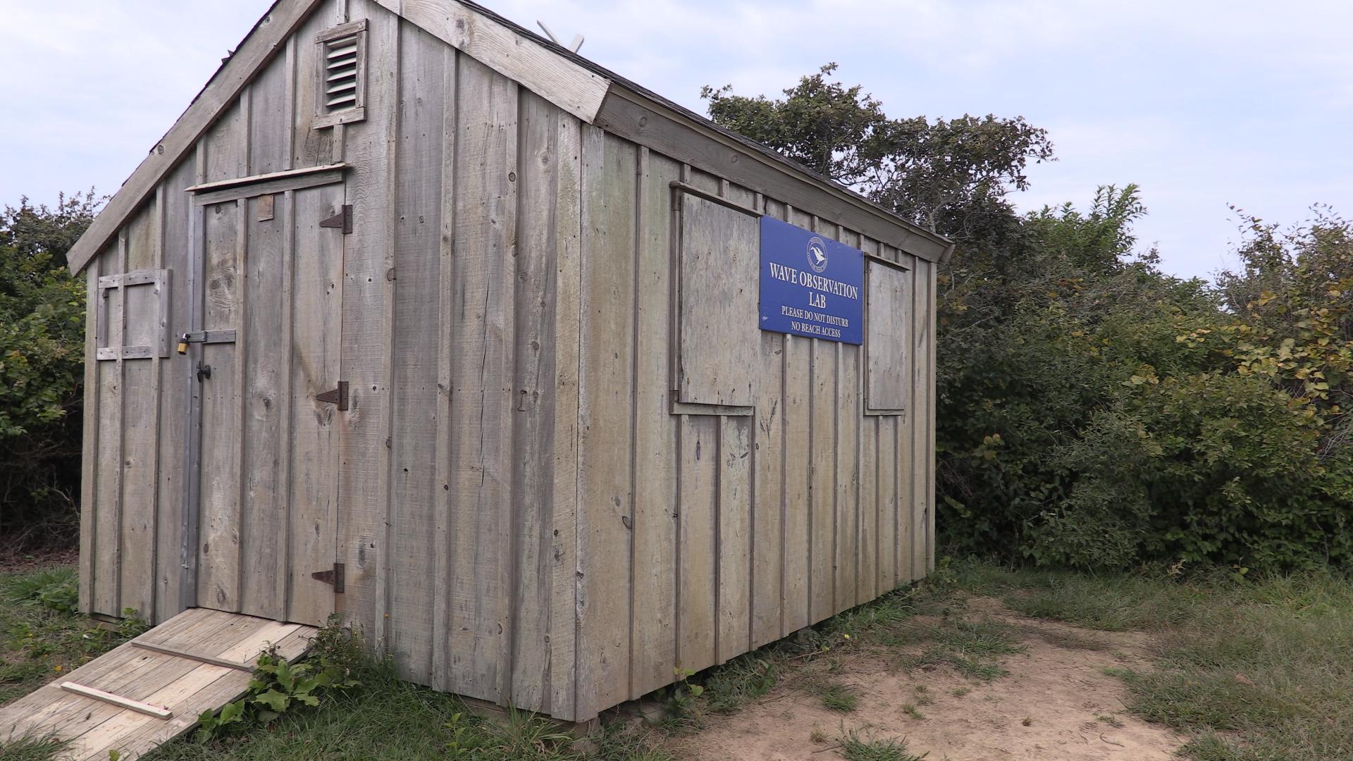 A wooden shed with a blue sign saying Cape Cod Wave Laboratory on a dirt cliff.