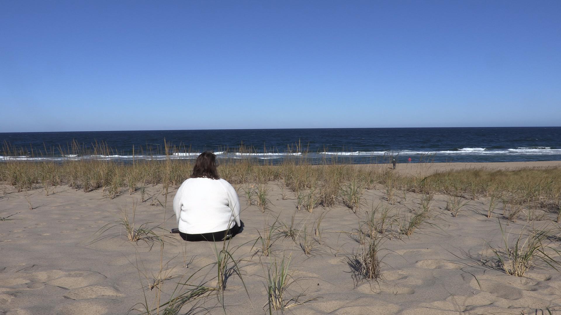A middle aged woman sitting on a sandy beach with her back to the camera on a sunny day. She appears to be looking at the ocean in front of her.