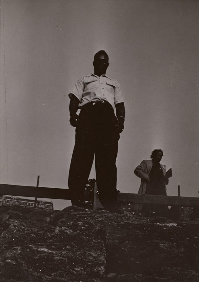 Black & white snapshot of a Black man in white shirtsleeves with a Black woman in the background. Photograph taken from a low vantage point