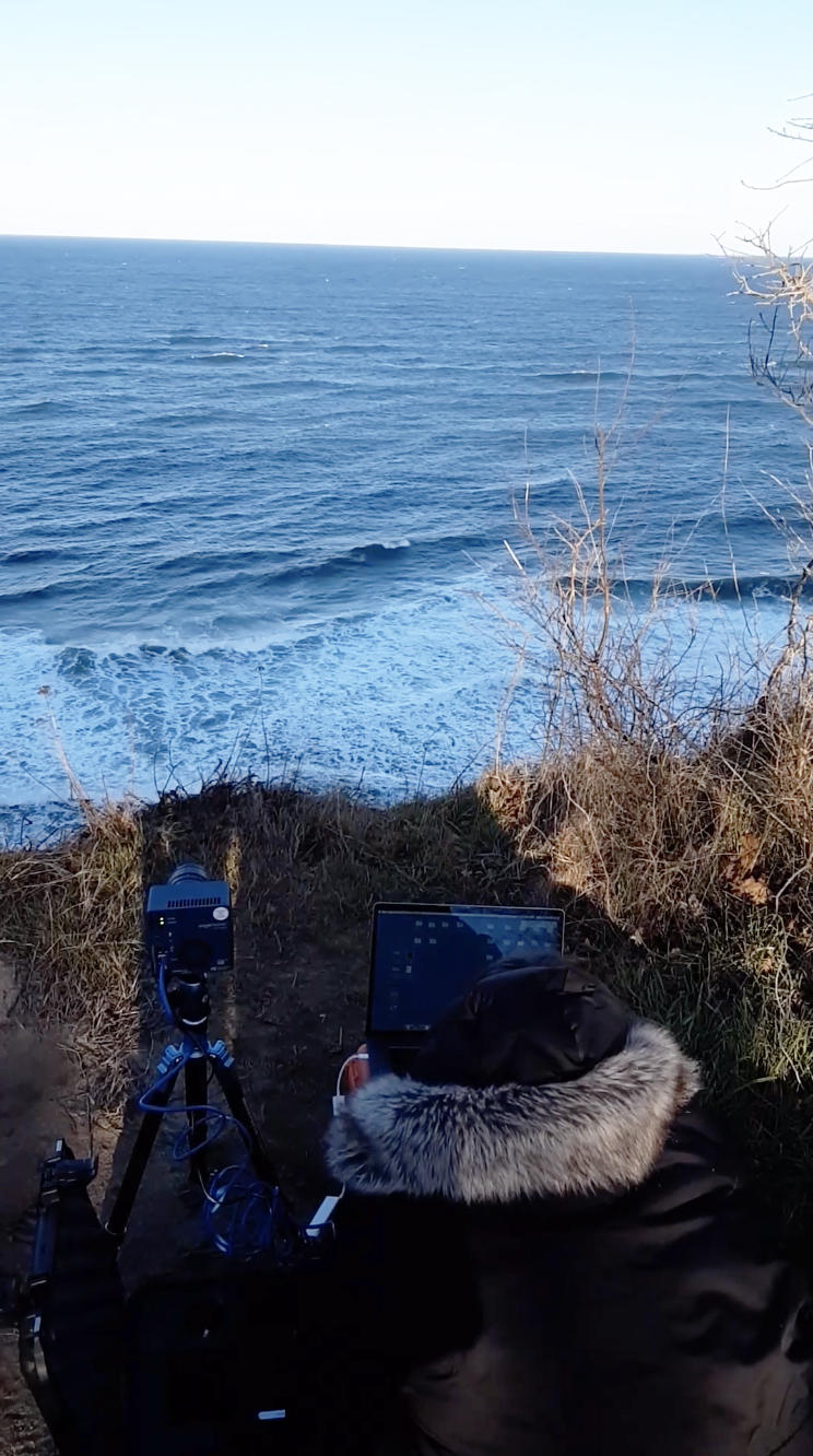 Person in heavy coat works with an Edgertronic Camera and laptop high above the sea.