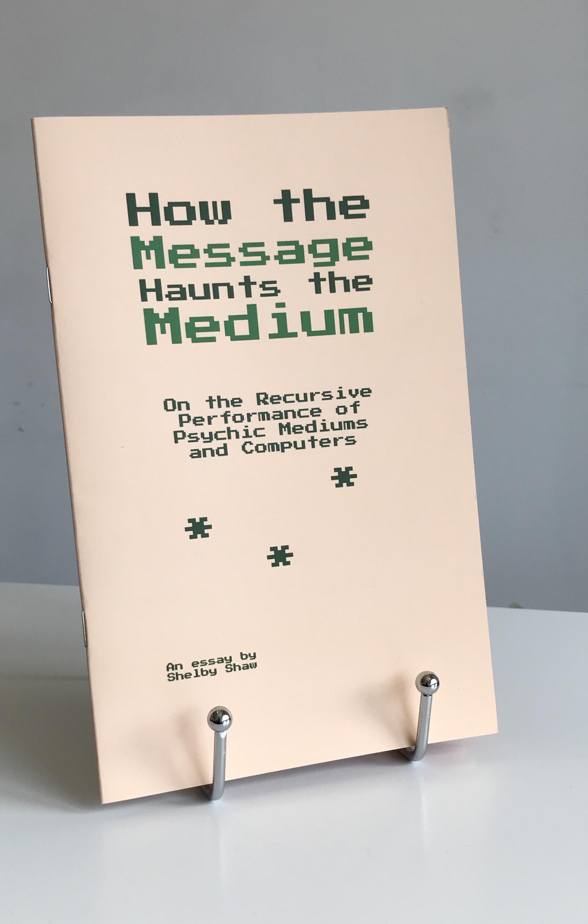 The book 'How the Message Haunts the Medium' which is peach colored with green font for the title and three large green asterisks underneath, is displayed on a silver book stand atop a white tabletop with a white wall in the background.