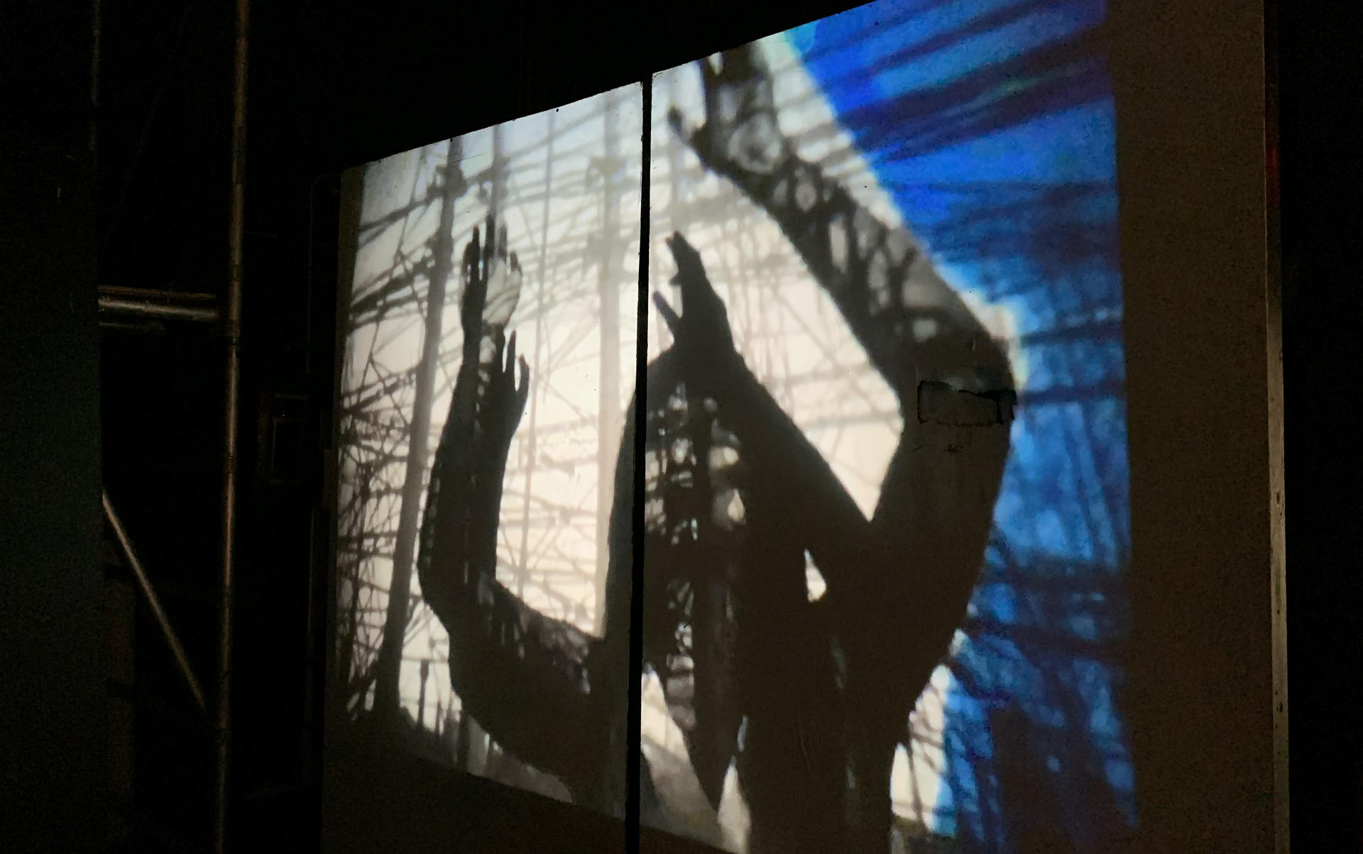 Frame from an animation lights up a dark room. It shows a human figure reaching amidst a web of lines like scaffolding or telephone poles and lines 