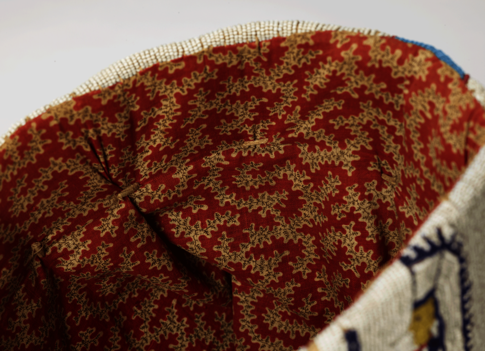 Interior detail of an object, covered in a dense print of cream flowers on a red background. The outer edge of the object is beaded in blue, white, and yellow.