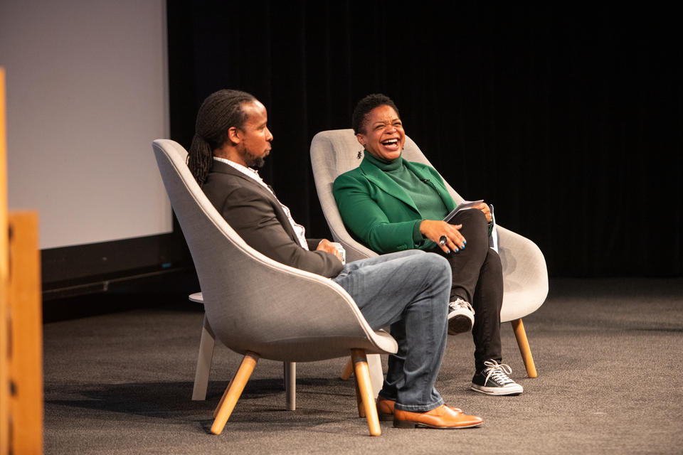 A Black man and woman sit on a stage. The woman, in a green blazer and turtleneck and black converse, laughs happily.