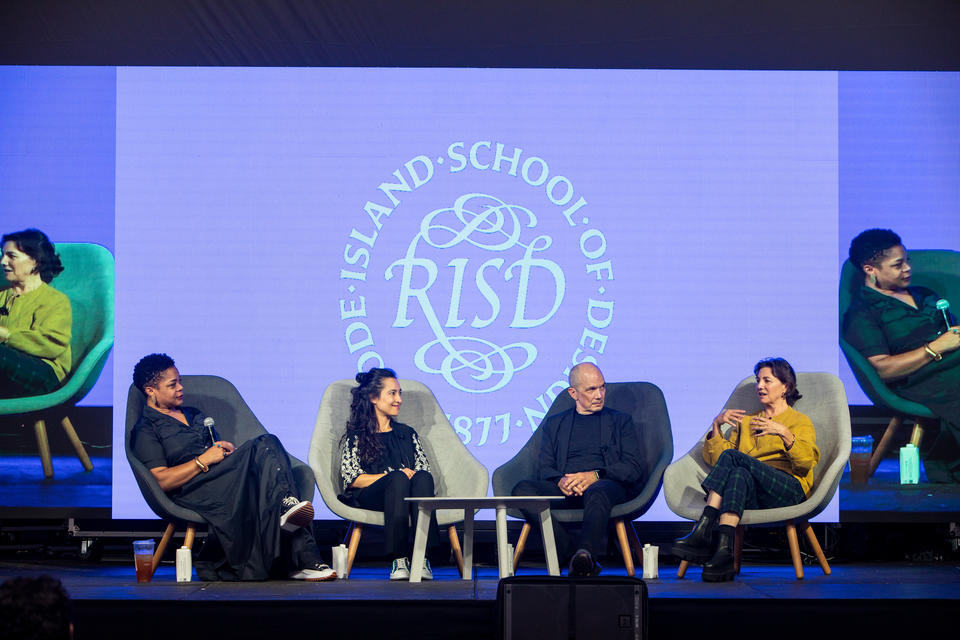 Four people sit in chairs on a stage. A white woman in a yellow sweater speaks while the others look on. The RISD seal is projected behind them.