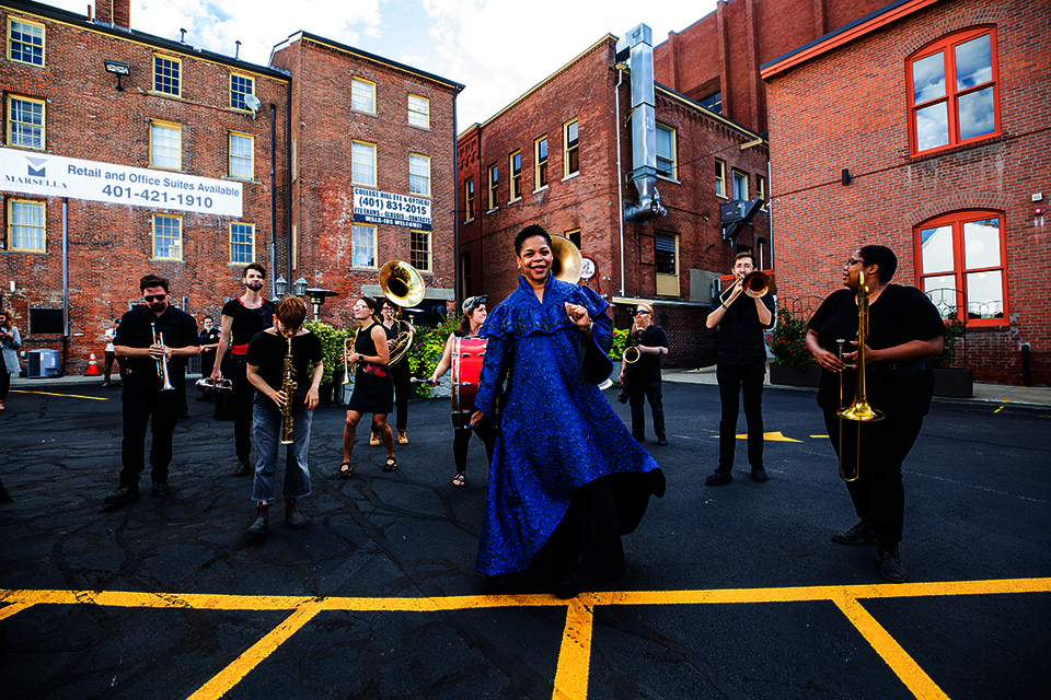 A Black woman in blue ceremonial robes dances in front of a small brass band, performing in a parking lot surrounded by brick buildings