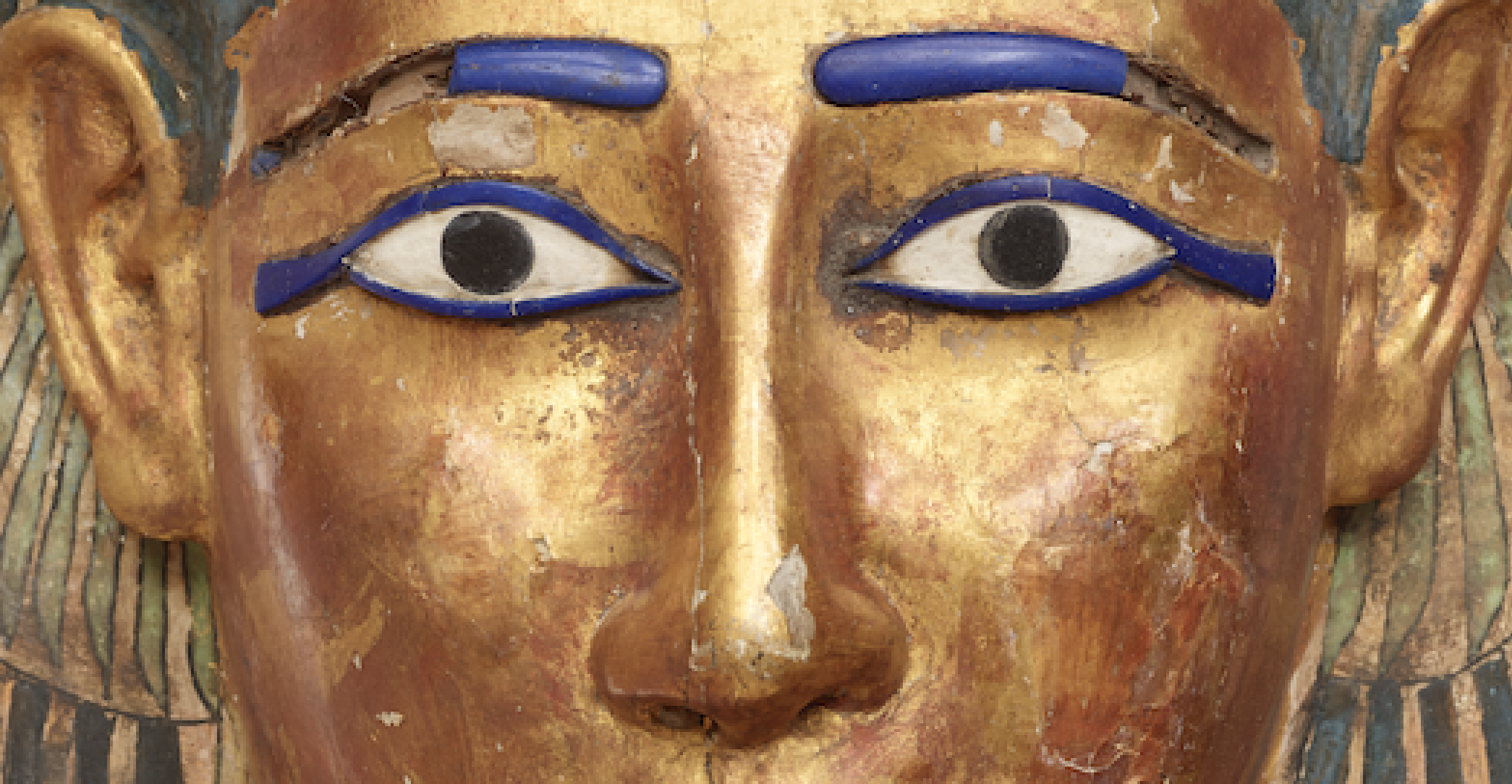 Close-up photo of Nesmin's gilded coffin, showing the face from the brows to the nose. The eyes are open and the eyebrows are a vivid inlaid blue.