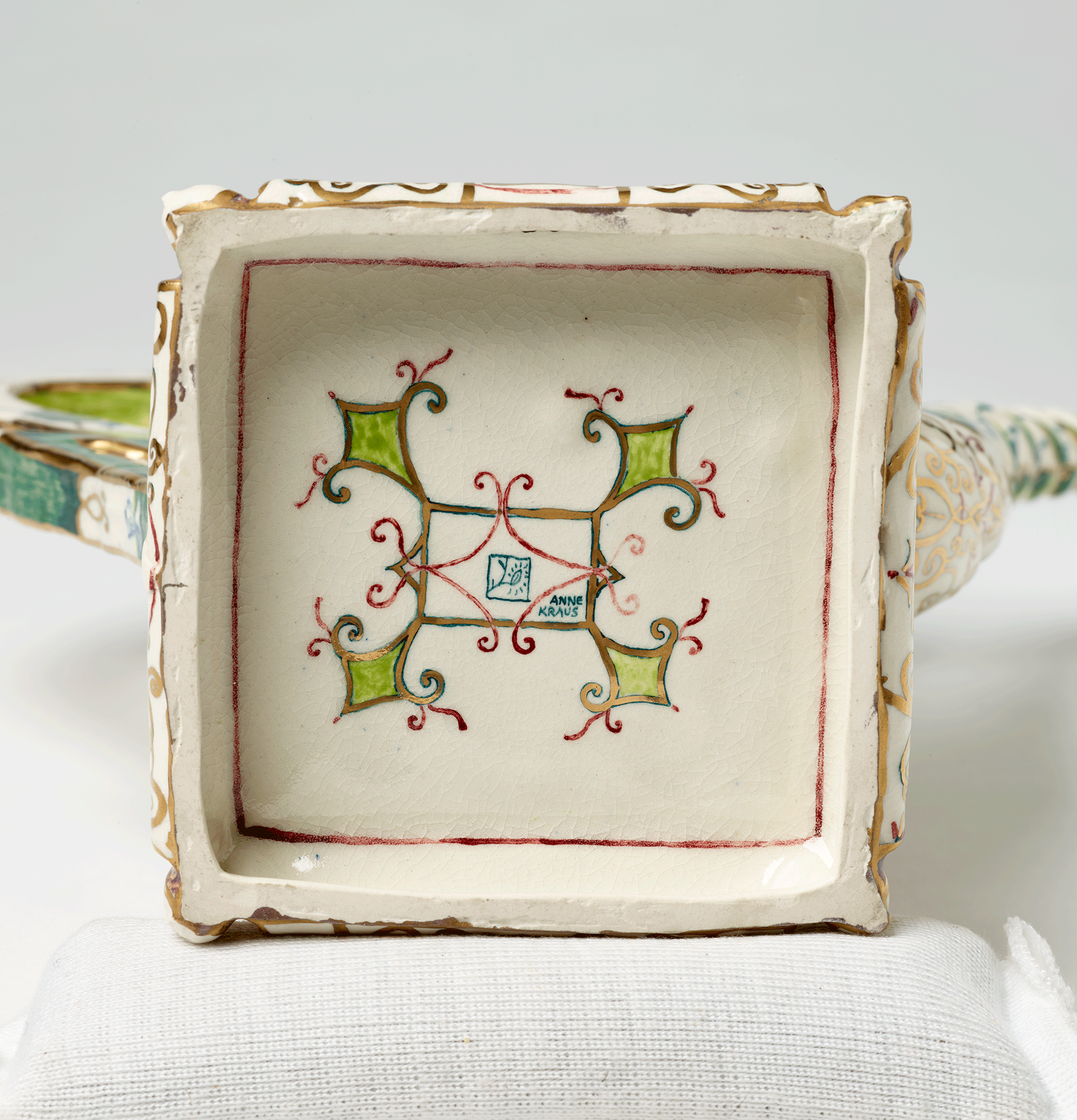 Square base of teapot with curving square and diamond-shaped painted design. The artist’s name, “Anne Kraus” is painted in small blue letters in the center square.