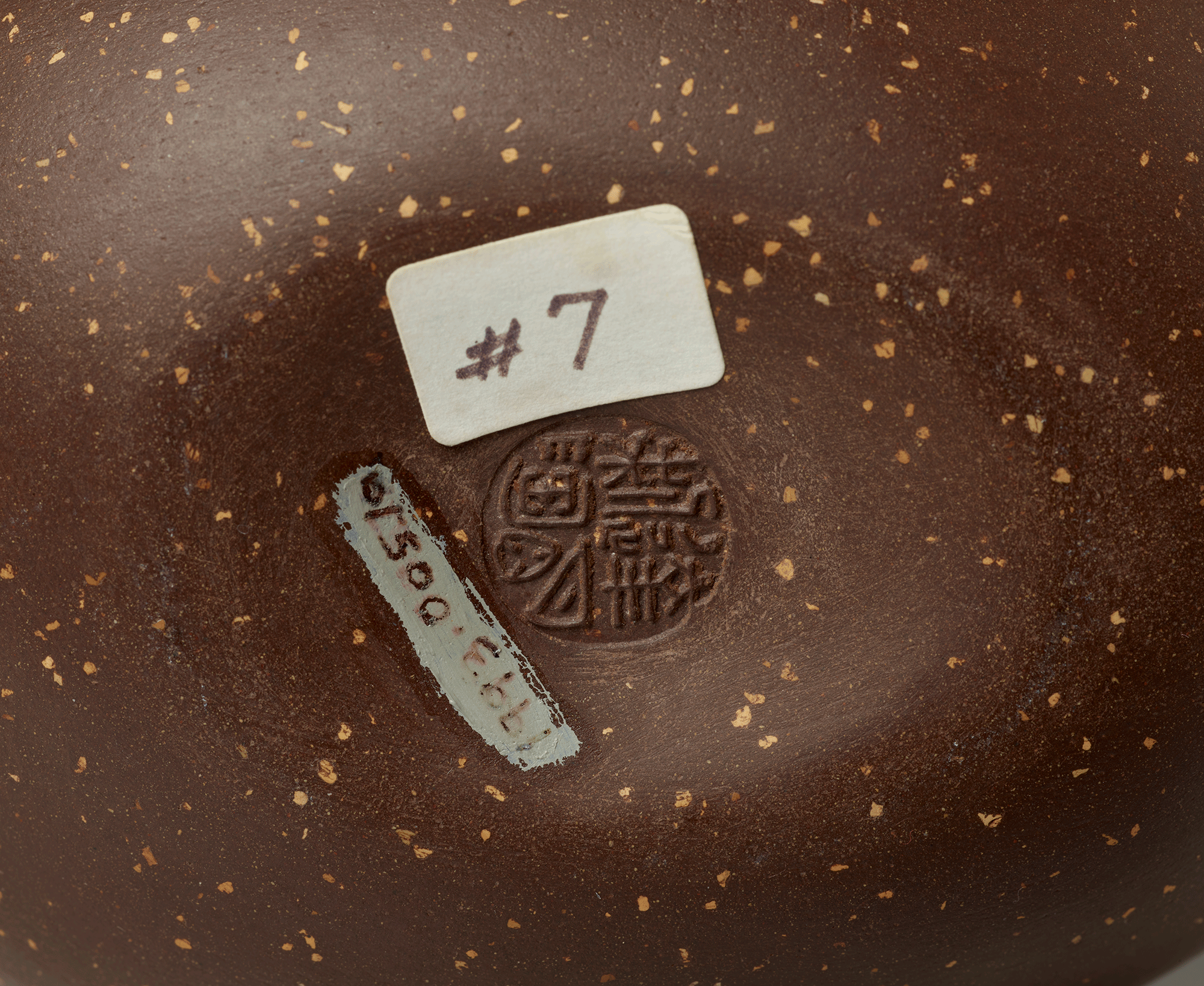 Underside of teapot. Chinese letters in a circular pattern stamped into the center. White, rectangular stick above with “# 7” written in brown ink. Accession number is visible.