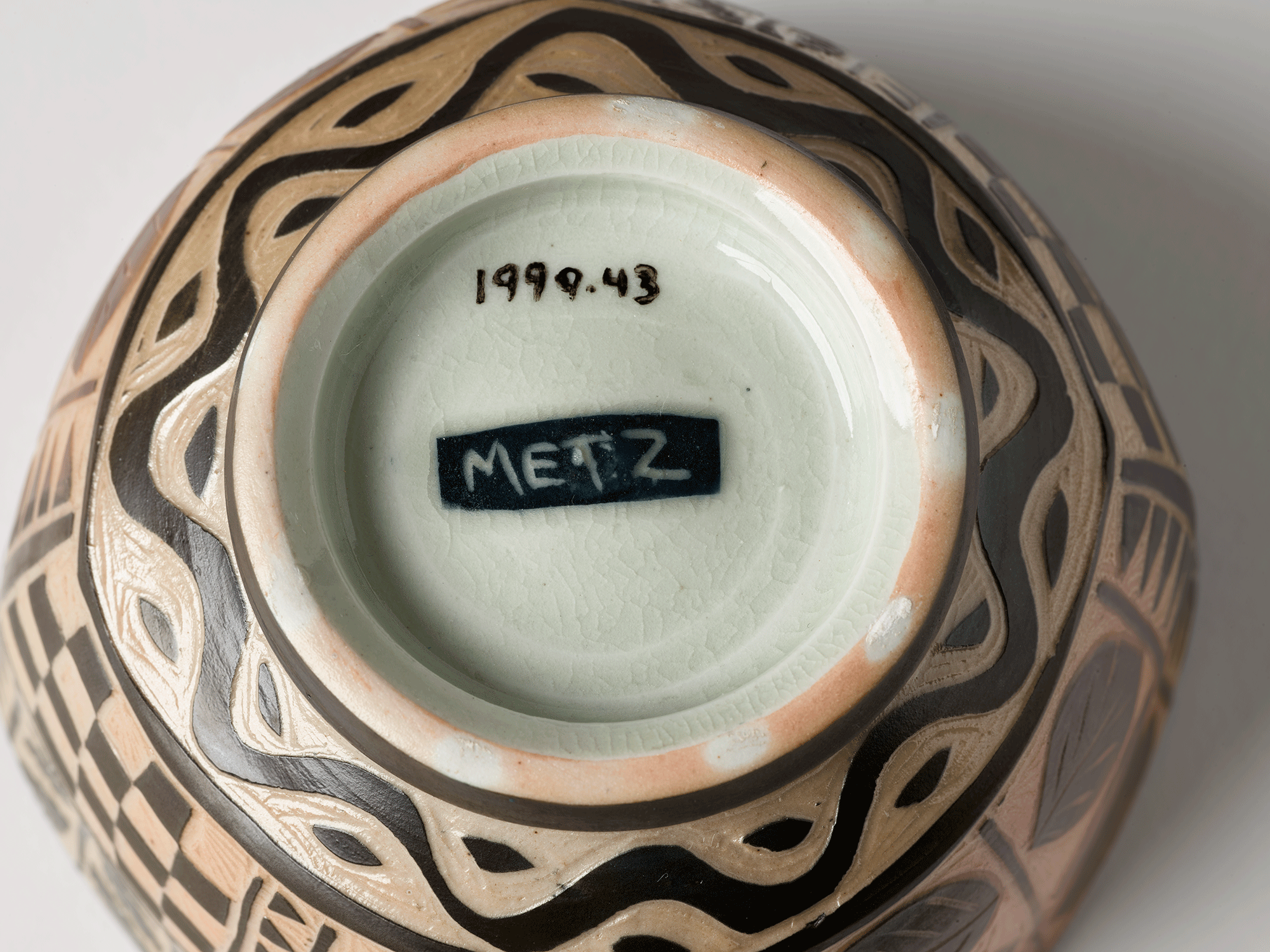 Underside of bowl. Wavy line and leaf-shaped patterning scratched into clay, circling the foot of the bowl. Light green interior of the foot has a black rectangle in the center with the name “Metz” painted inside. Accession number is visible.