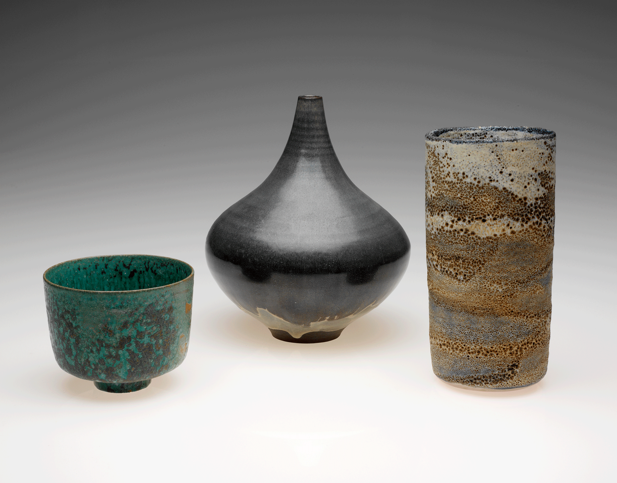 Group of three vessels. A small, green bowl is on the left. A dark gray bottle is in the middle, and a cylindrical vessel is on the right.