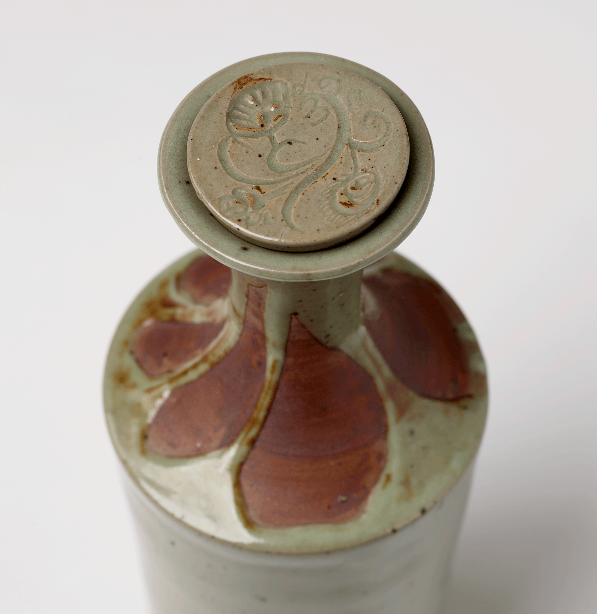 Detail of the top of the decanter. Rust colored petal shapes are carved into light green clay ground. Curving flower design on bottle lid.