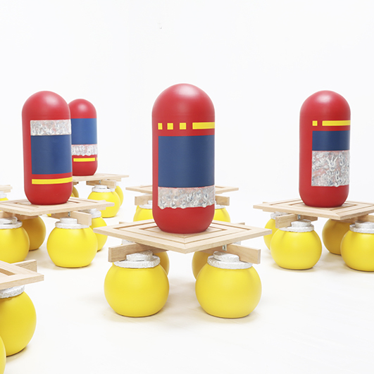 Color photograph; four bright red pill-shaped ceramic objects with blue, yellow, and silver rectangles painted on. They each sit on little square wooden platforms held aloft by four yellow spheres