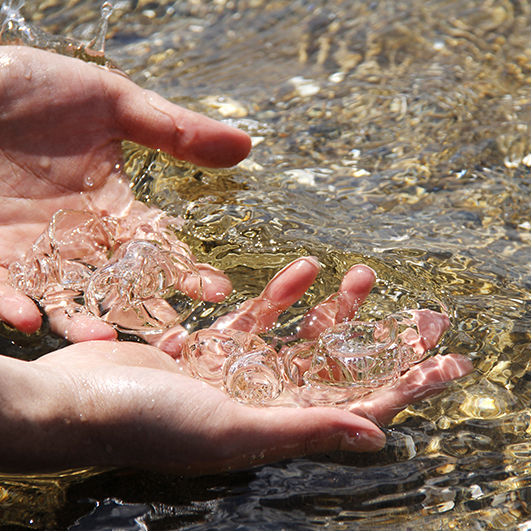 color photograph of a pair of hands submerged in crystal clear running water. The hands hold clear blobs of glass, which initially camouflage with the water.