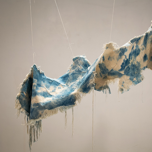 color photograph of a piece of canvas suspended from the ceiling. Its edges are frayed and it has cyan colored splotches