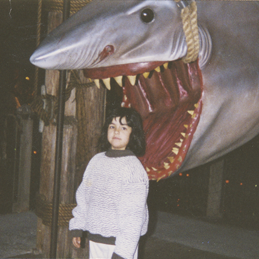 Color photograph of a young white girl with shoulder length dark hair, wearing a light colored bulky sweater, standing beneath a threatening shark sculpture. The shark looks like it will bite her head off.