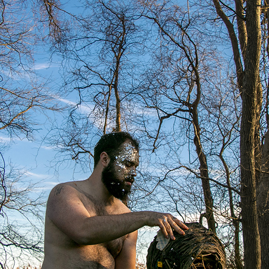 Color photograph of a shirtless, bearded man sitting outside amongst leafless trees. He holds a basket and large silver glitter covers his face