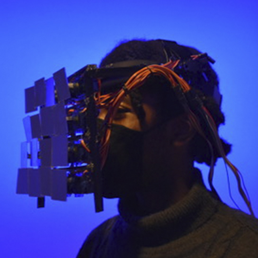 Color photo of a young black person wearing a wearable mechanical sculpture over their face.