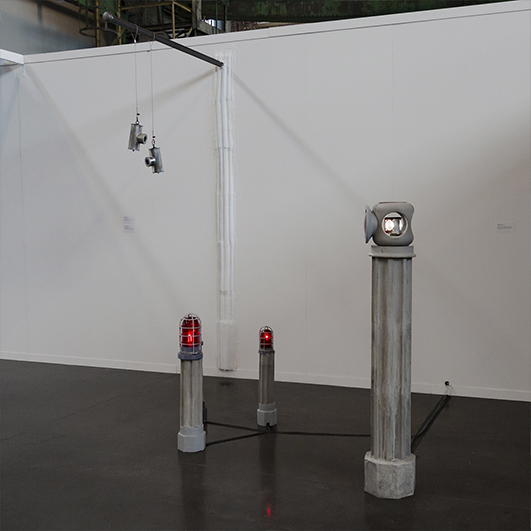 color photo of three grey columns with red lanterns at the their tops and a larger column in the foreground with a white lantern in a gallery setting
