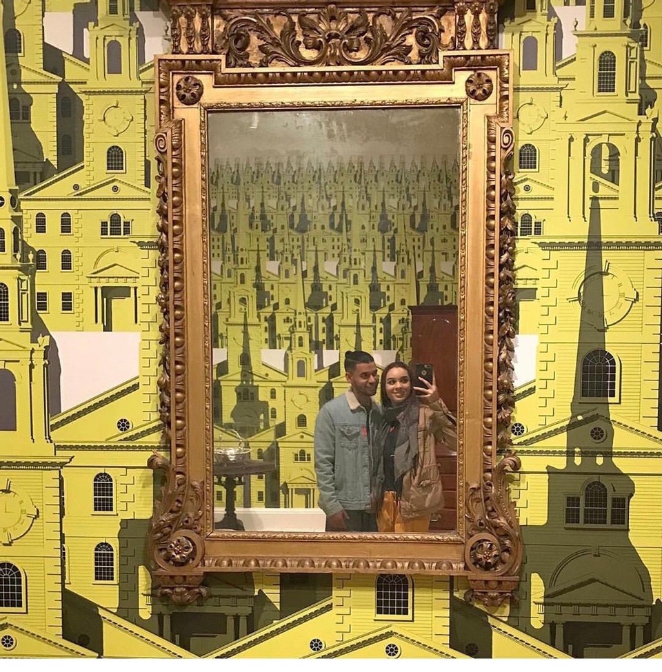 /Two%20smiling%20young%20people%20take%20a%20selfie%20in%20an%20ornate%20antique%20mirror.%20On%20the%20wall%20behind%20the%20mirror%20is%20bright%20yellow%20wallpaper%20patterned%20with%20architectural%20images.