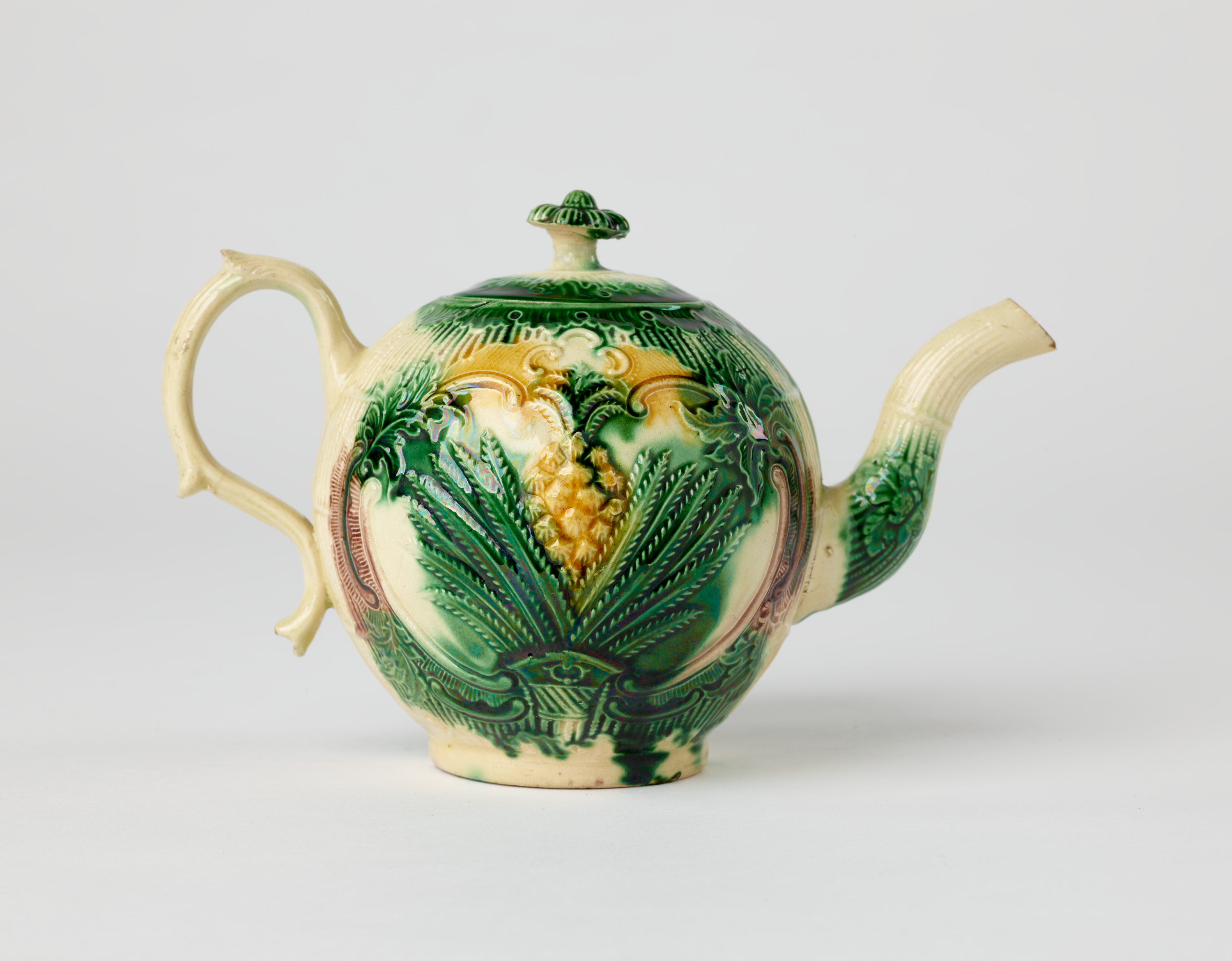 /A%20bulbous%20ceramic%20teapot%20with%20a%20long%20curved%20spout%20and%20handle%2C%20white%2C%20dark%20green.%20and%20yellow%20glaze%2C%20and%20floral%20decorations%20ranging%20from%20leaves%20to%20a%20more%20central%20pineapple.