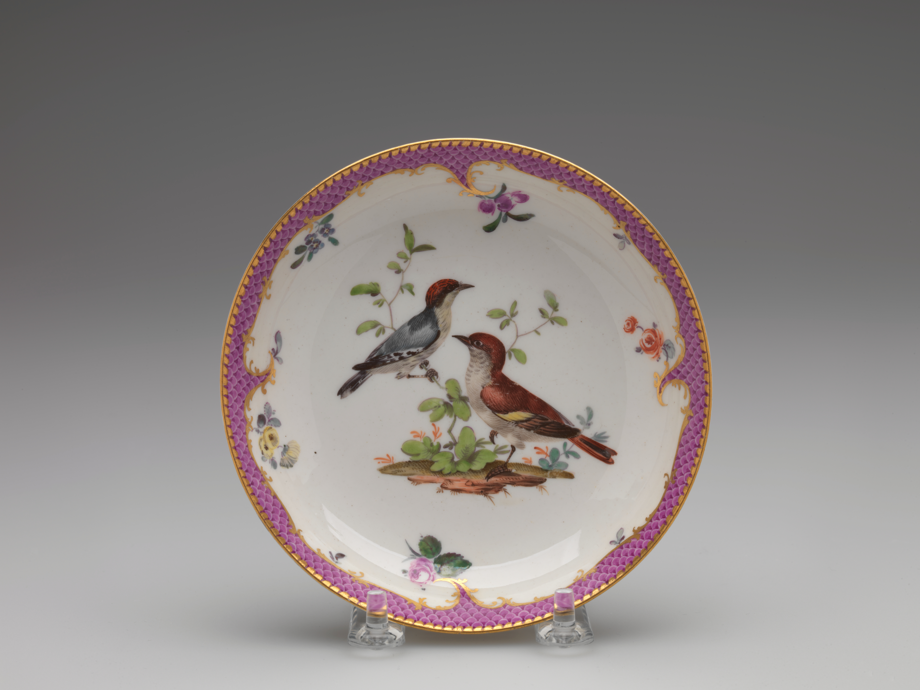 /A%20white%20saucer%20with%20gilded%2C%20pink%2C%20floral%20decorations%2C%20and%20two%20birds%3B%20one%20bird%20is%20blue%20with%20a%20red%20head%20positioned%20on%20a%20branch%2C%20the%20other%20is%20standing%20on%20the%20ground.