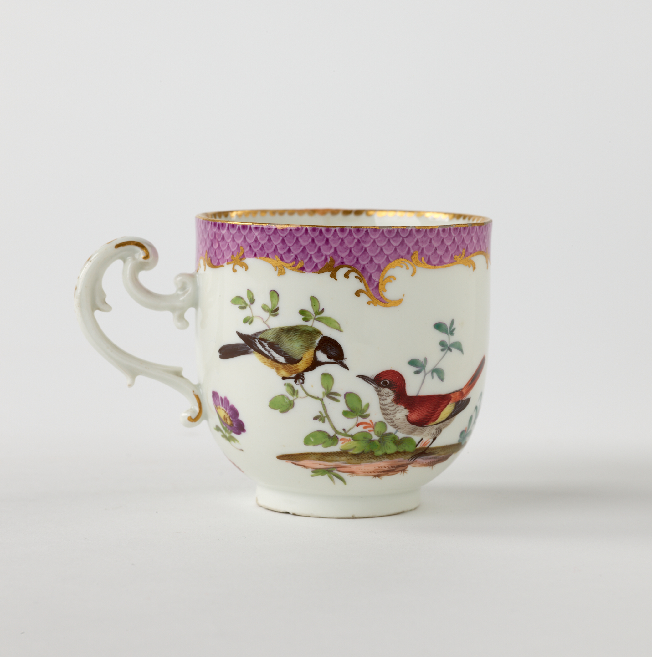 /A%20white%20teacup%20with%20a%20handle%20that%20has%20swirls%2C%20gilding%2C%20pink%2C%20and%20floral%20decorations.%20There%20are%20two%20birds%2C%20one%20gray%20and%20yellow%2C%20the%20other%20is%20larger%20and%20mainly%20red.