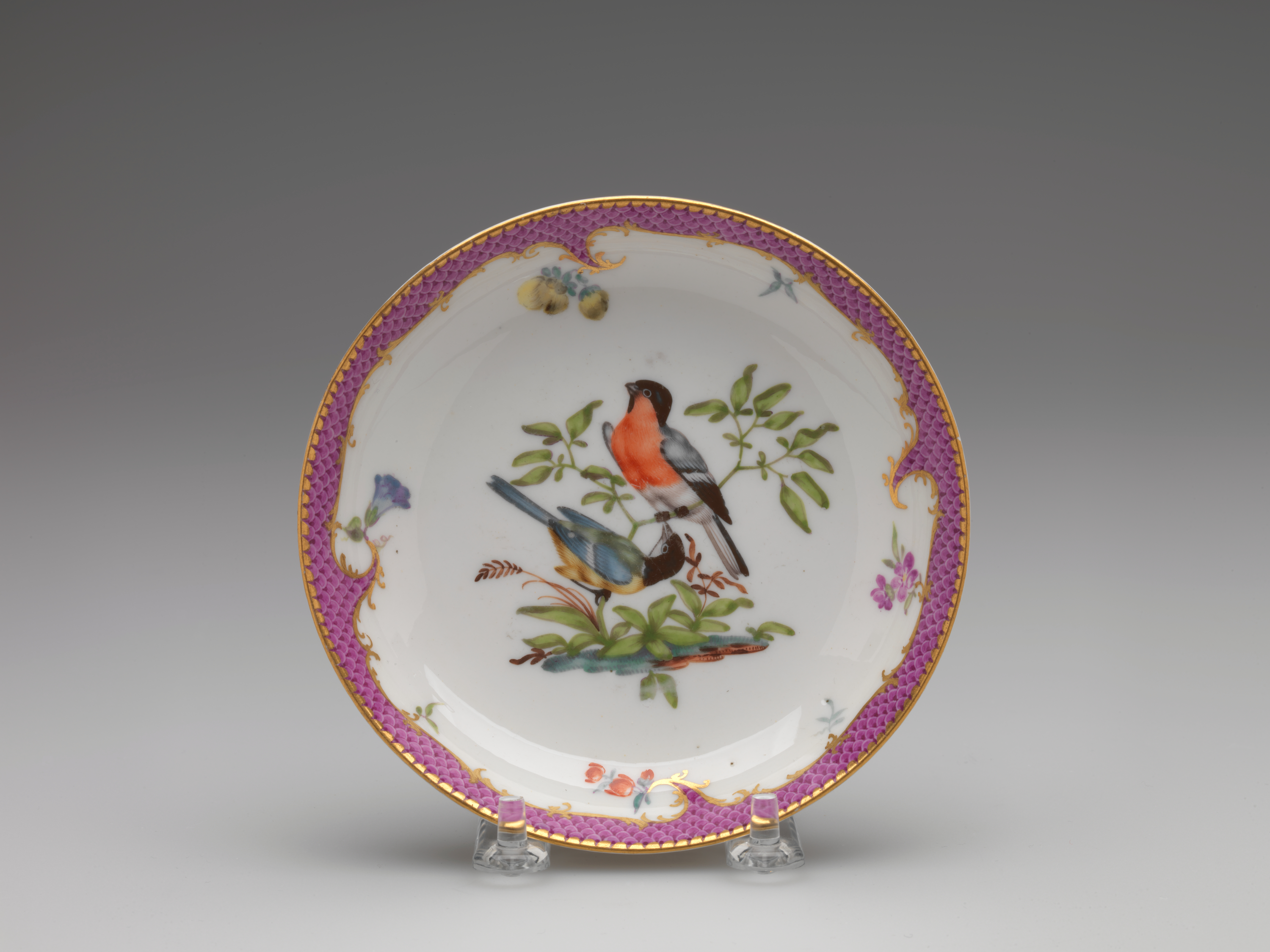 /A%20white%20ceramic%20plate%20with%20a%20gilded%20and%20pink%20edge%2C%20floral%20patterns%2C%20with%20two%20birds%20surrounded%20by%20floral%20elements%2C%20positioned%20on%20branches%2C%20are%20blue%2C%20white%2C%20black%2C%20red%2C%20and%20yellow.