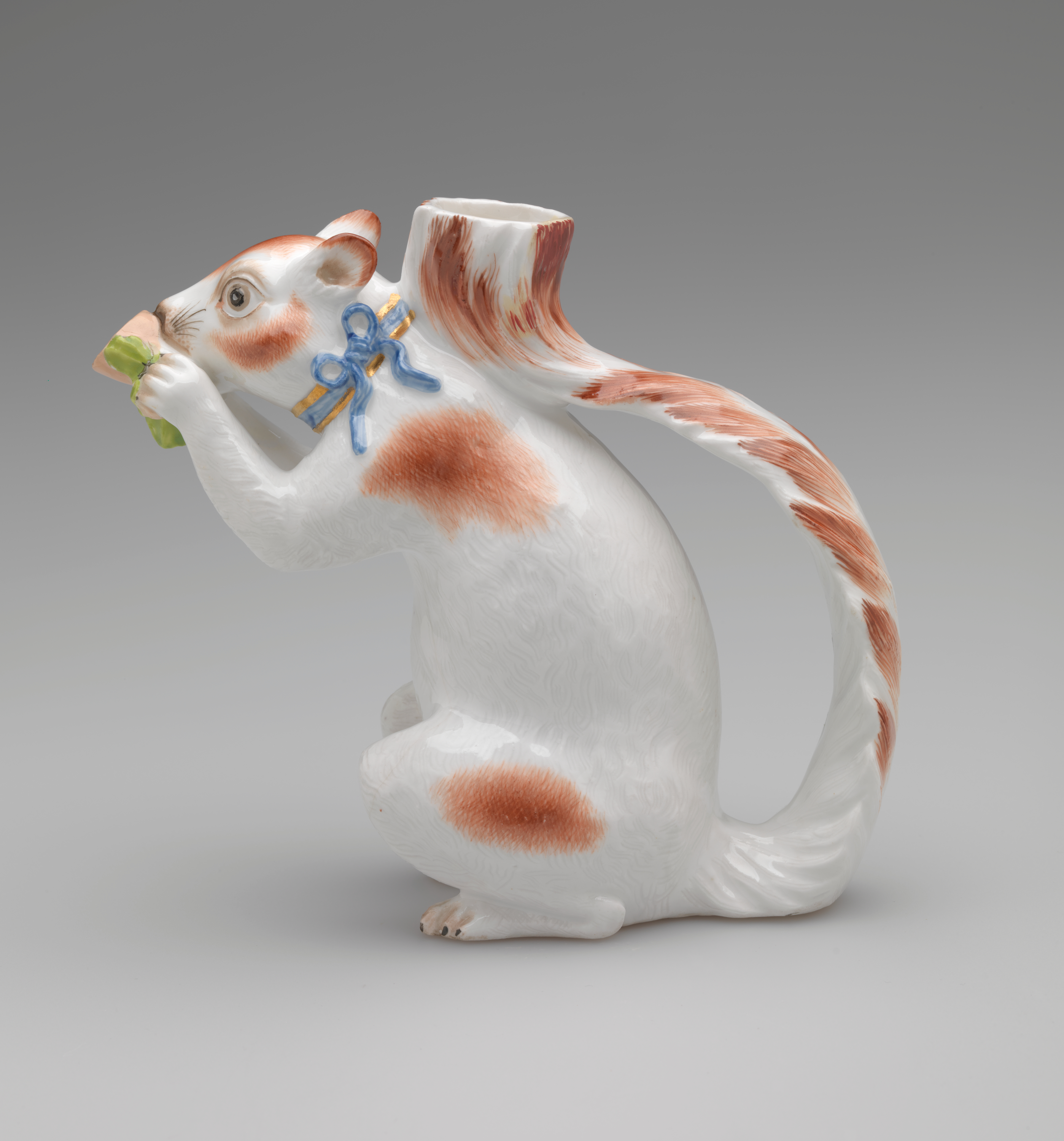 /Vessel%20in%20the%20shape%20of%20a%20white%20squirrel%20with%20brown%20patches%20and%20a%20blue%20ribbon%20arounds%20its%20neck.%20It%20holds%20a%20brown%20nut%20with%20green%20leaves%20up%20to%20its%20face.