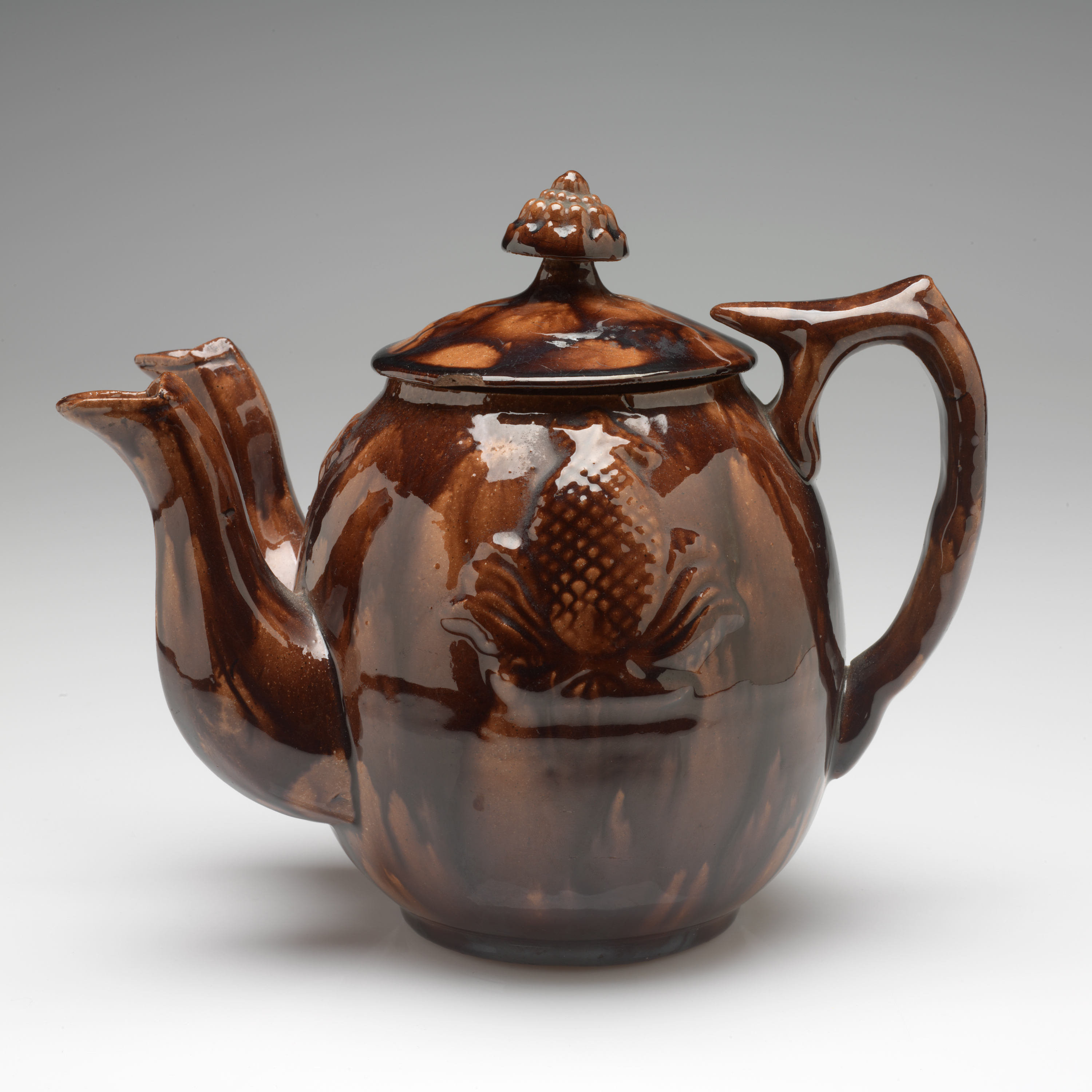 /A%20mottled%20brown%20teapot%20with%20an%20image%20of%20a%20pineapple%20in%20the%20center.%20Two%20spouts%20are%20on%20the%20left%20side%2C%20and%20a%20rounded%20handle%20with%20a%20flat%20top%20faces%20right.