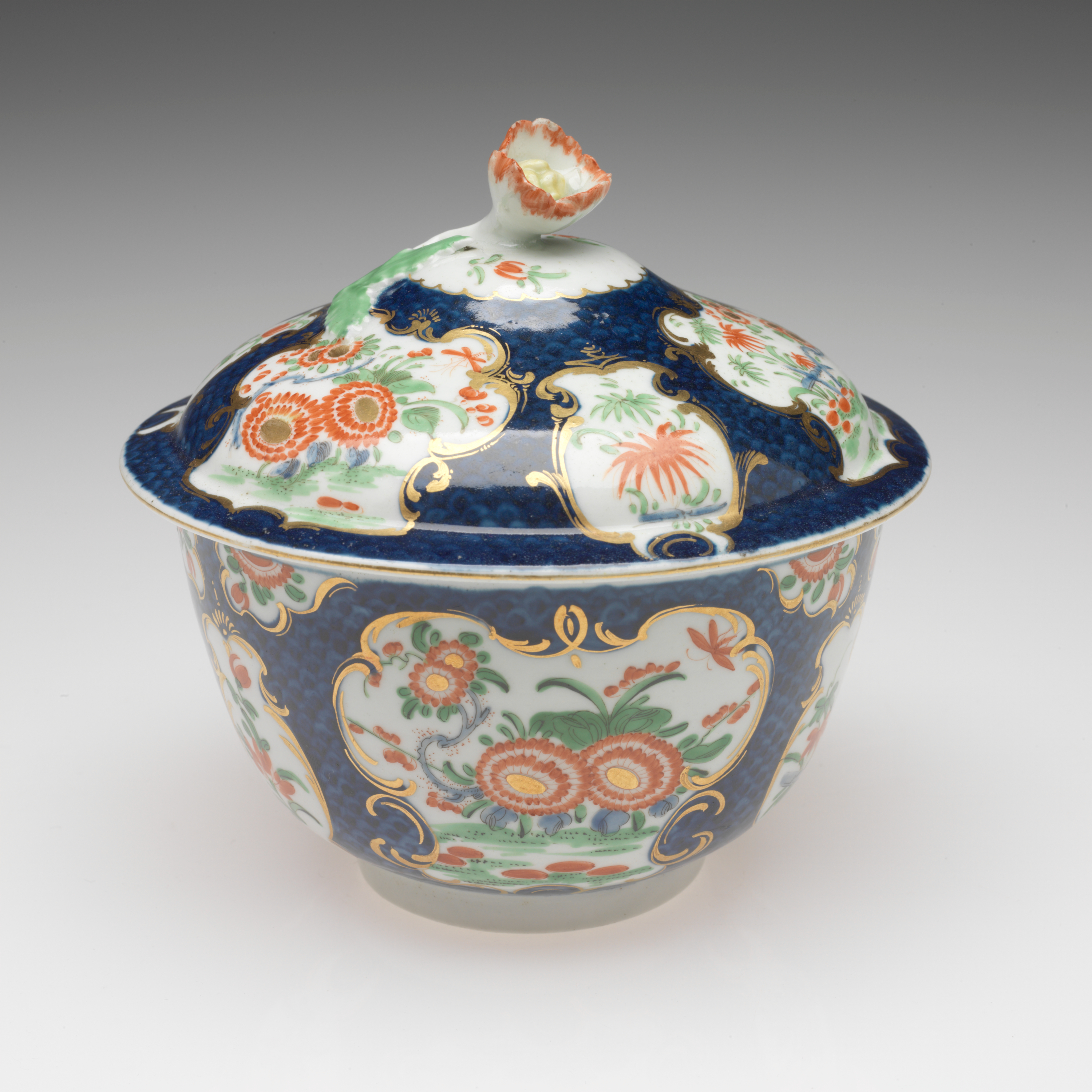 /A%20lidded%20sugar%20bowl%20with%20floral%20decorations%20in%20gilded%20vignettes%2C%20surrounded%20by%20dark%20blue%20glaze%2C%20the%20finial%20on%20the%20lid%20is%20a%20sculptural%20red%2C%20white%2C%20and%20yellow%20flower.%20