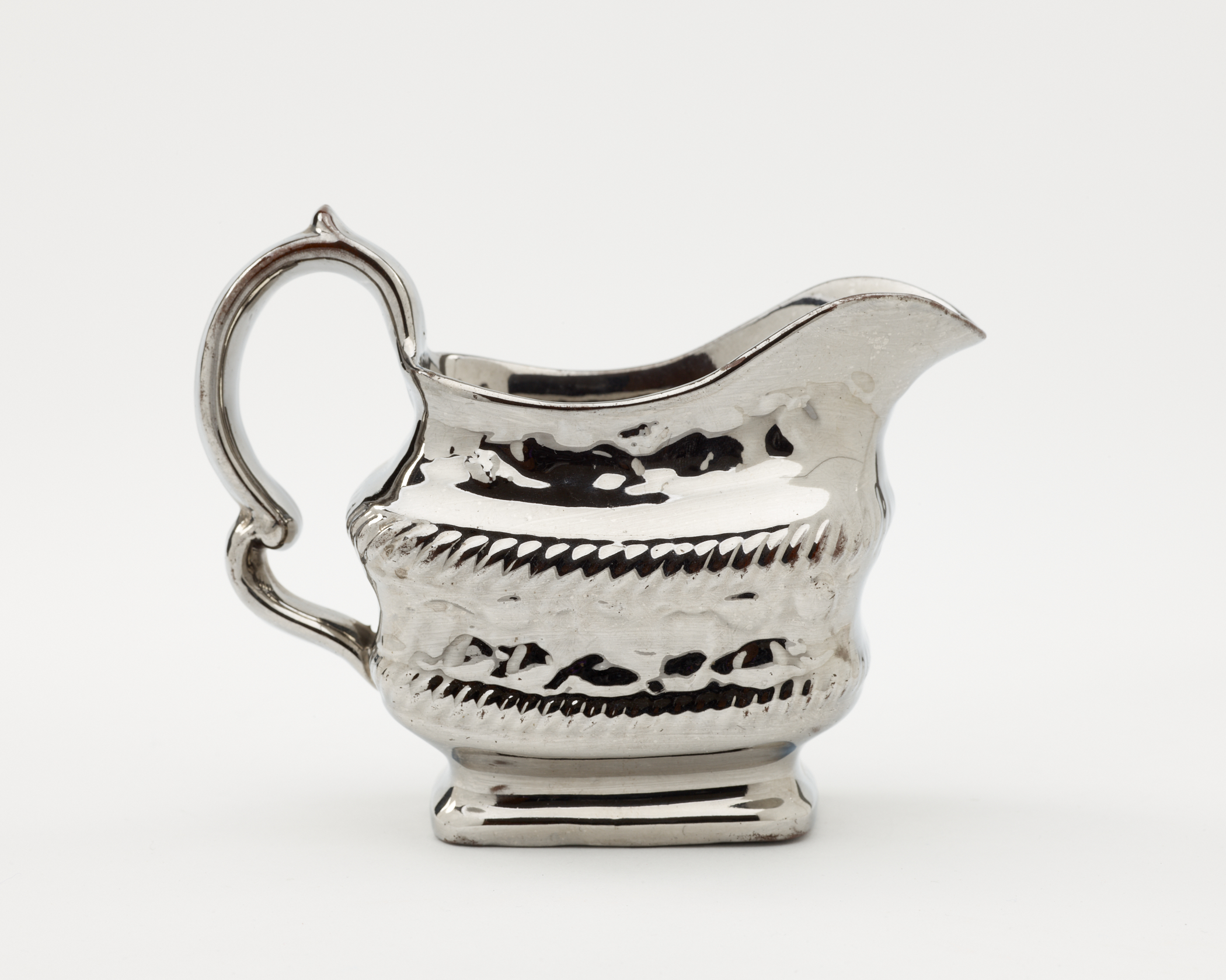 /A%20silver%20luster%20creamer%20with%20a%20decorative%20handle%2C%20a%20rounded%20square%20body%20with%20sculptural%20decorations.
