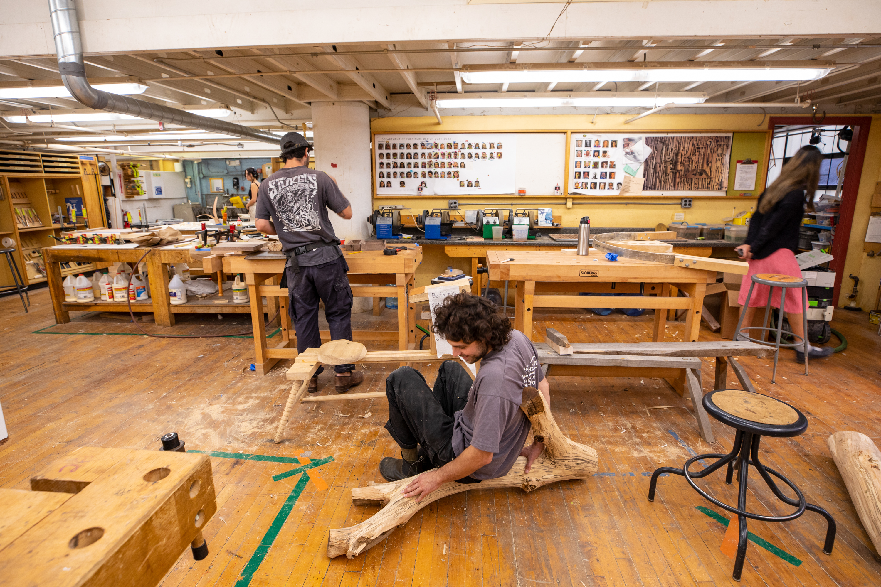 /Three%20students%20in%20a%20woodshop.%20One%20works%20at%20a%20bench%2C%20one%20sits%20on%20a%20piece%20of%20driftwood%2C%20and%20the%20third%20walks%20around.