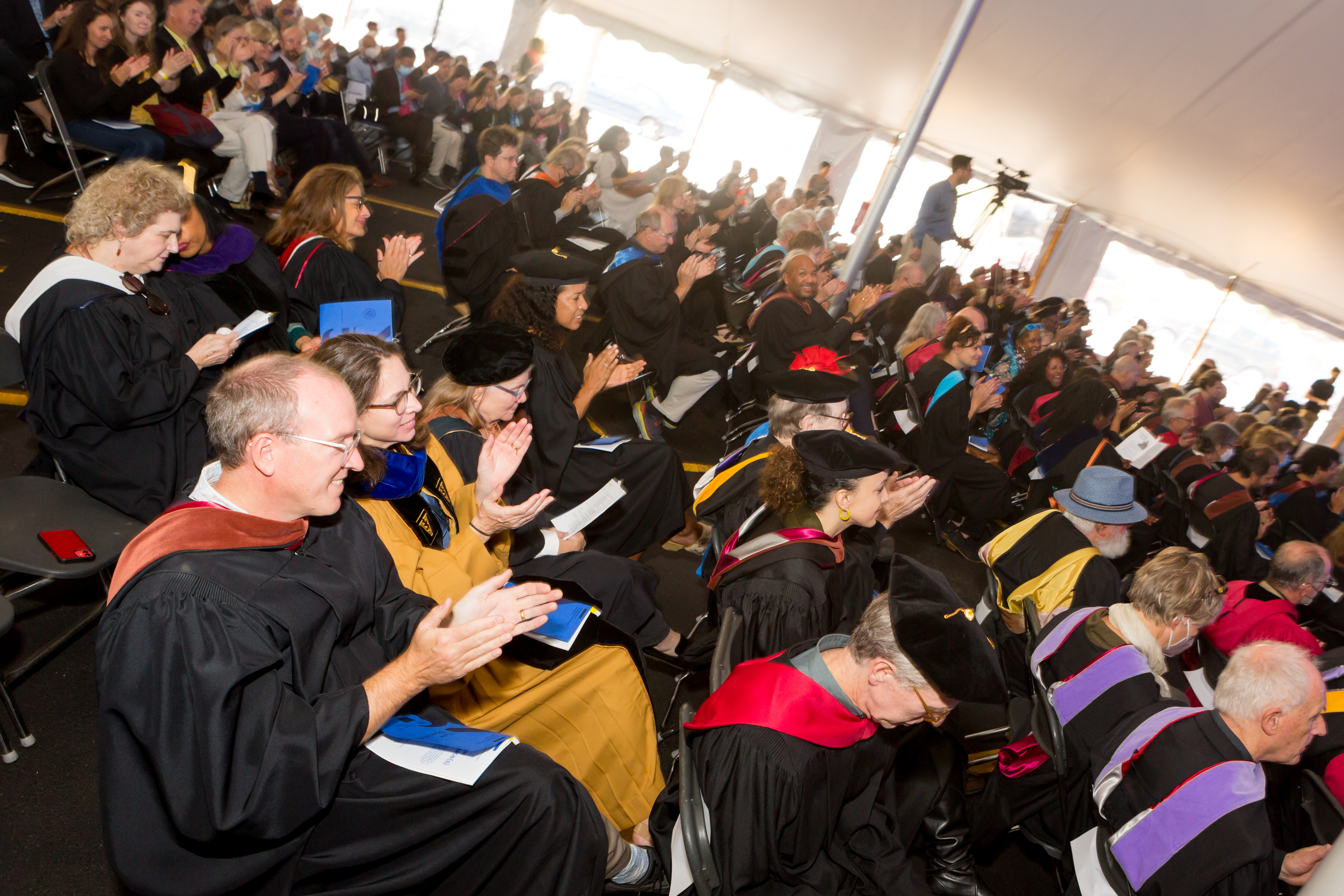 /A%20crowd%20of%20seated%20people%20dressed%20in%20masters%20and%20doctoral%20robes%20applaud.
