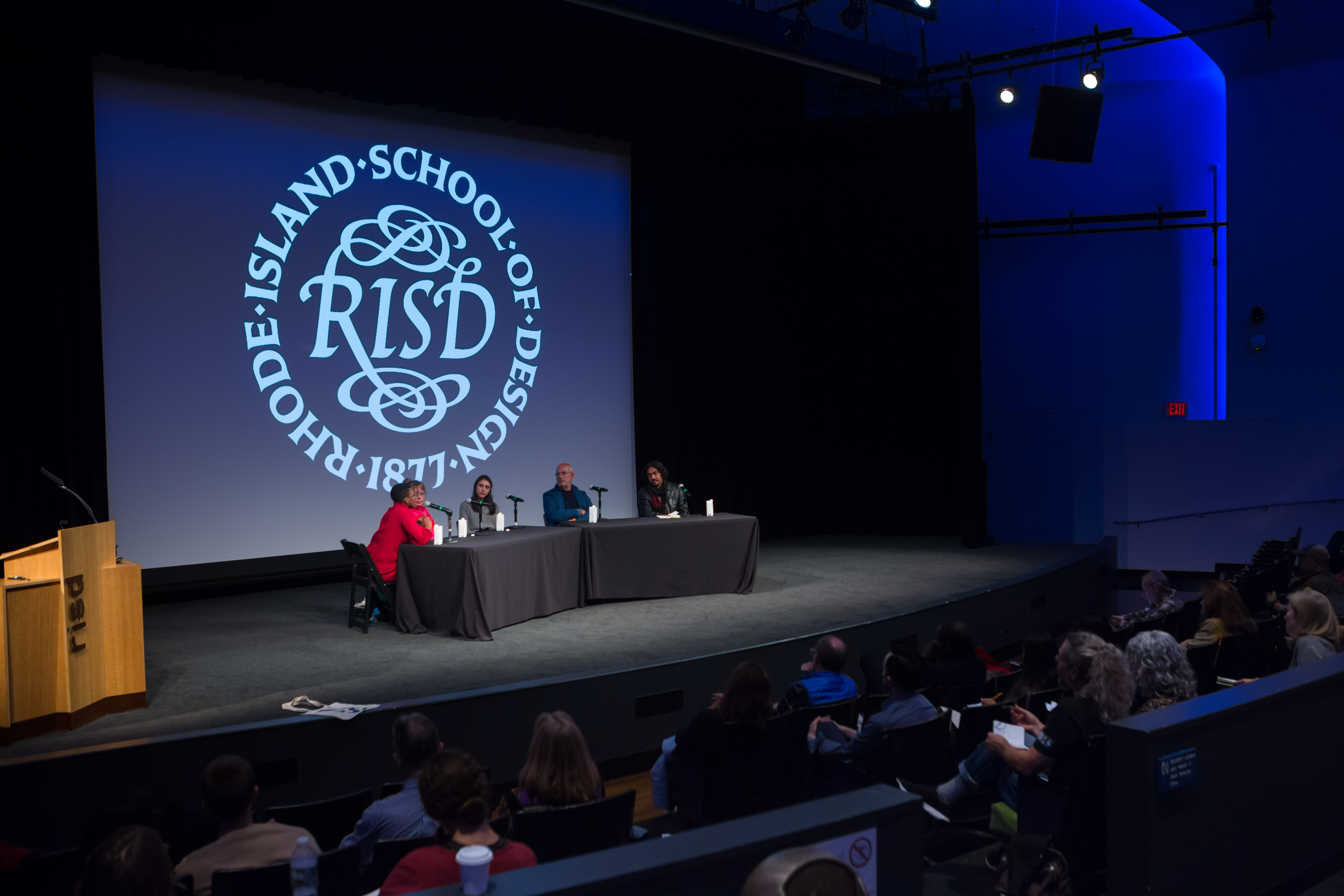/Five%20panelists%20sit%20at%20a%20table%20onstage%20in%20an%20auditorium.%20The%20RISD%20seal%20is%20projected%20behind%20them.