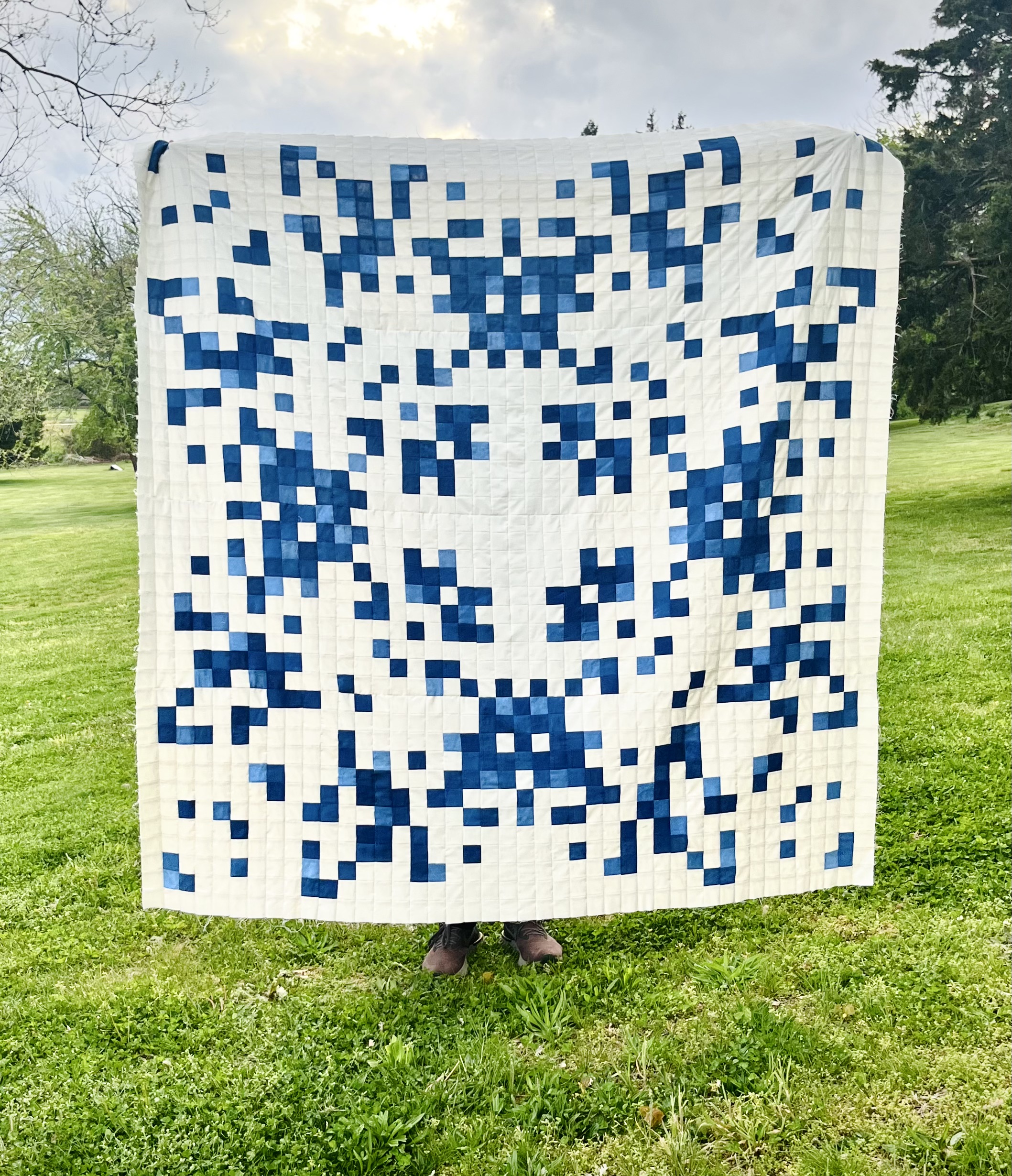 /A%20large%20blue%20and%20white%20quilt%20is%20held%20up%20outside%20in%20a%20field%20of%20green%20grass%20at%20golden%20hour.