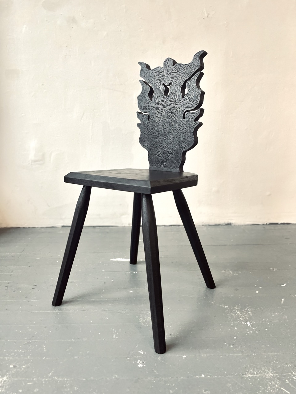 /A%20black%20chair%20stands%20on%20a%20wooden%20floor.%20The%20base%20of%20the%20chair%20is%20simple%2C%20utilitarian%20and%20the%20back%20is%20intricately%20carved.