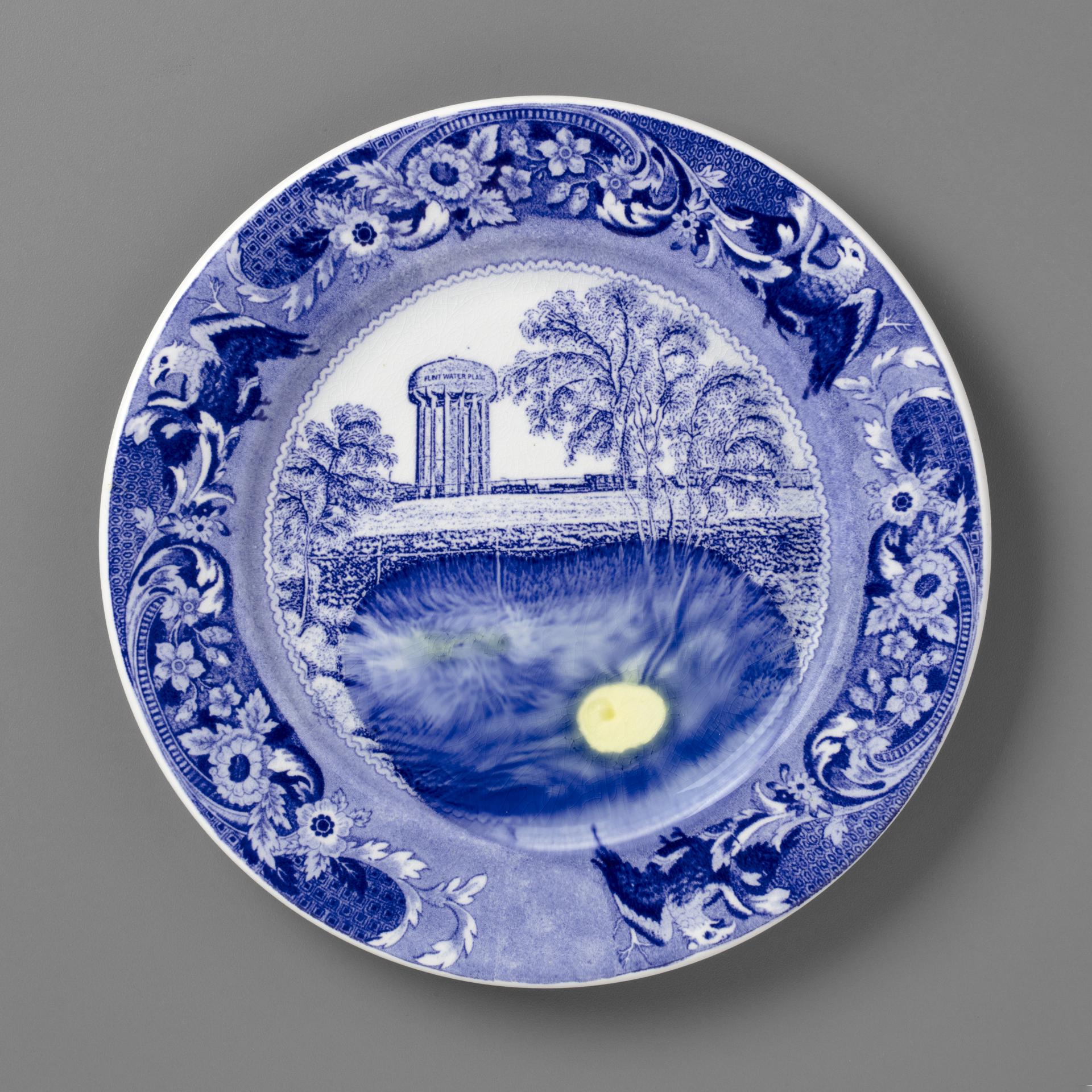 blue and white transferware plate depicting trees and a water tower labeled Flint, with lead melted onto the plate