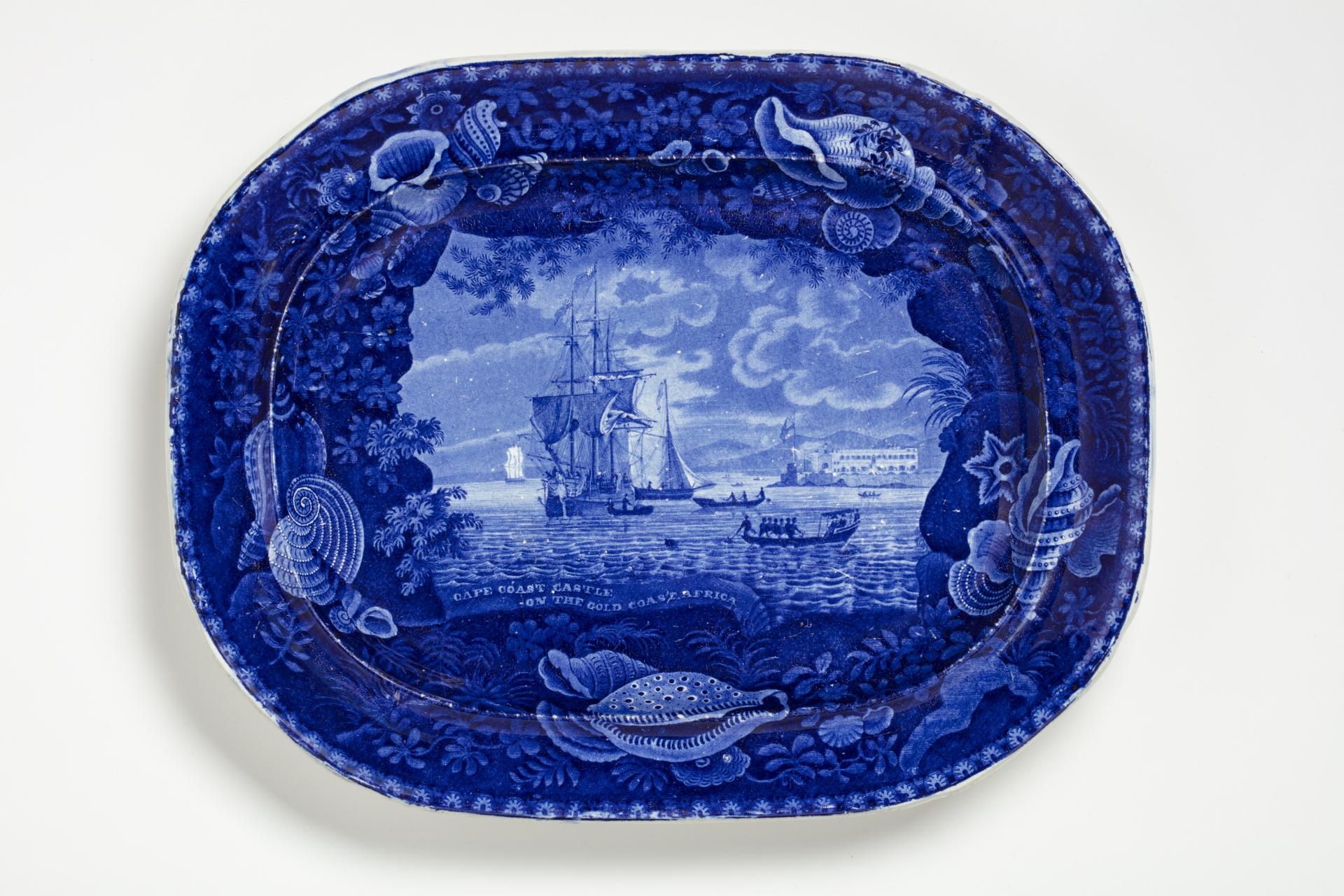 blue and white transferware platter depicting a slave ship off near the Cape Coast Castle, with a decorative shell border