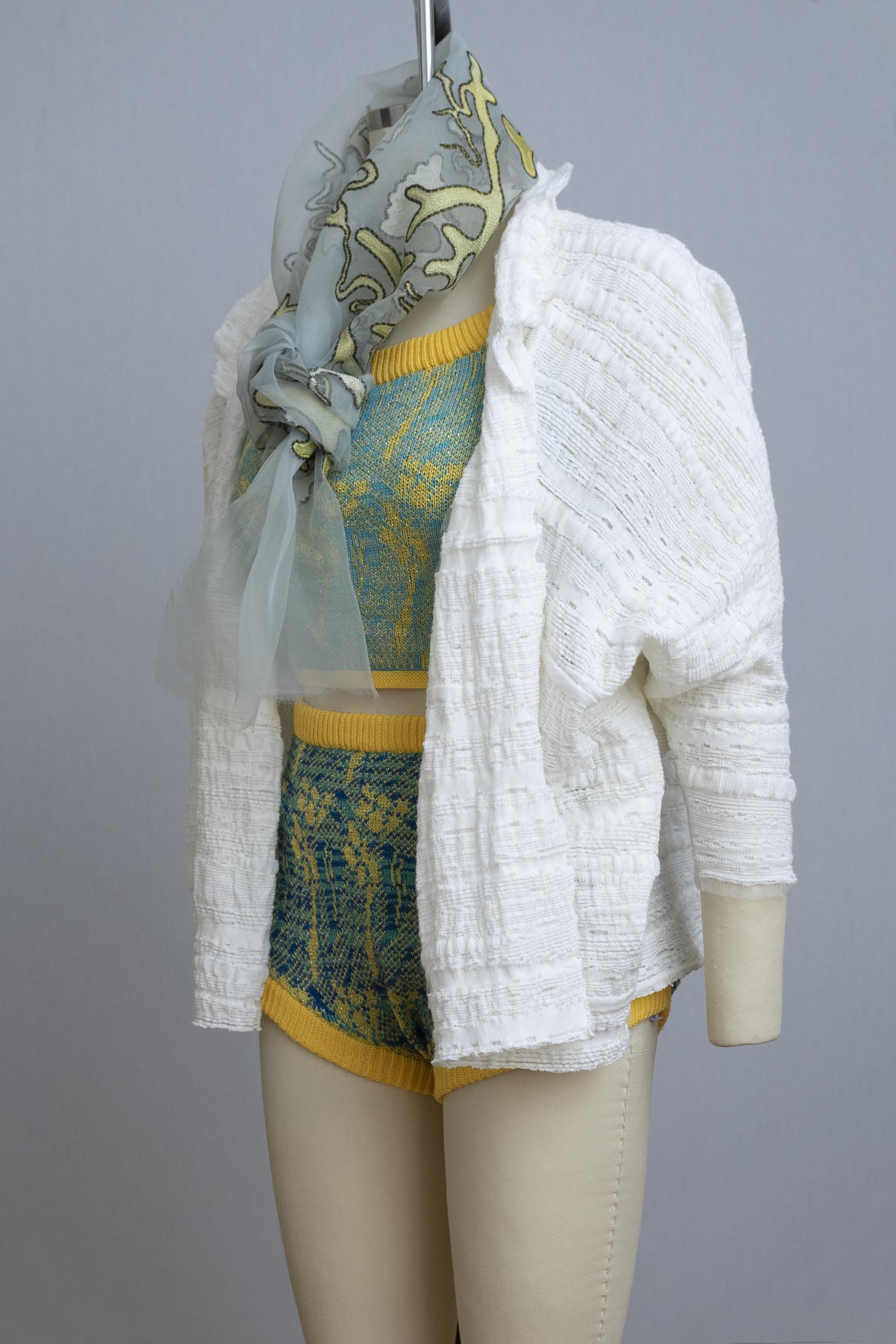 dress form wearing a knit playsuit in blues and yellows, a white knit sweater, and a sheer painted scarf