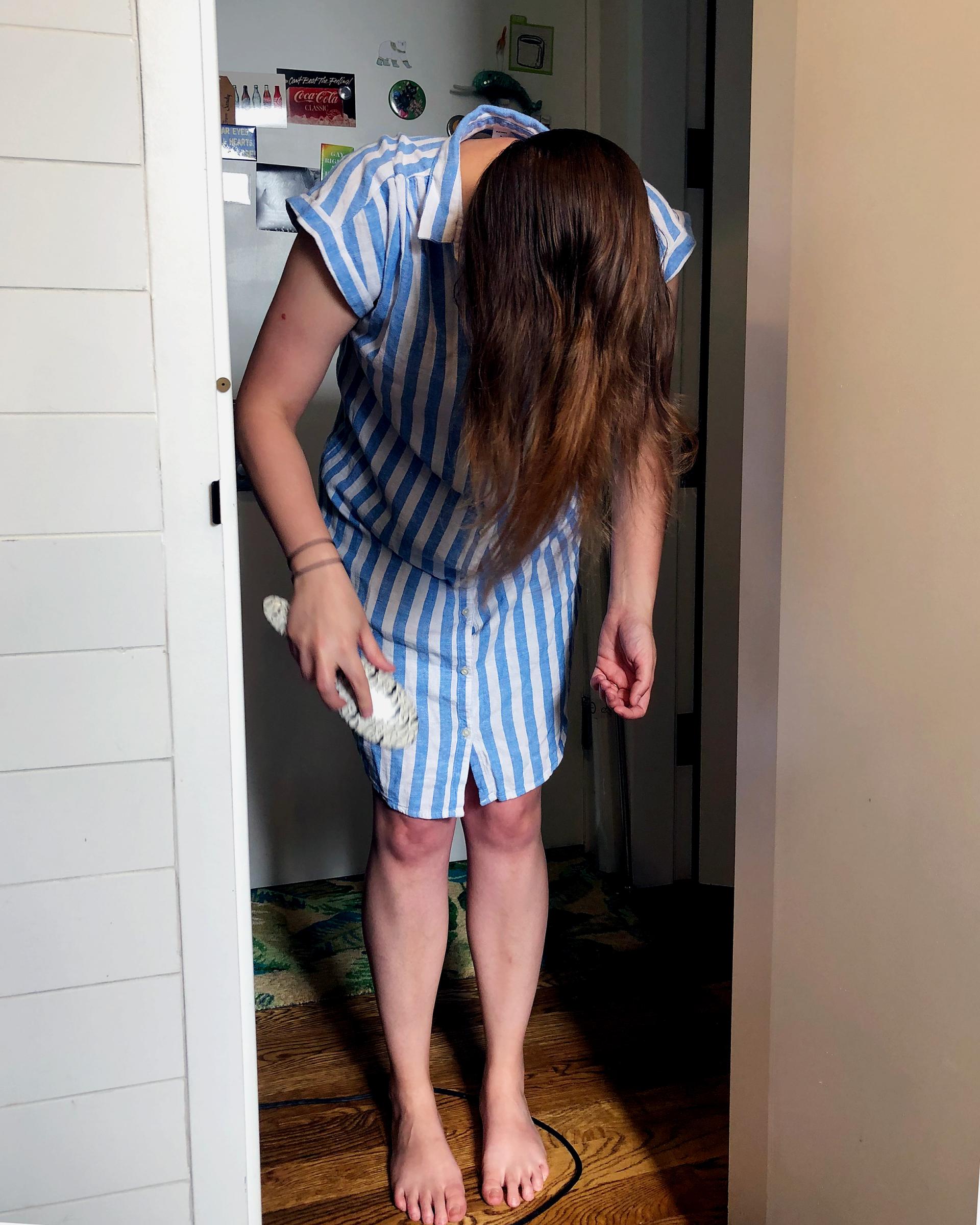 A photograph of a white feminine person in a doorframe. They are barefoot, wearing a blue and white striped shirt-dress and holding a hairbrush, having just pulled it through their long brown hair. 