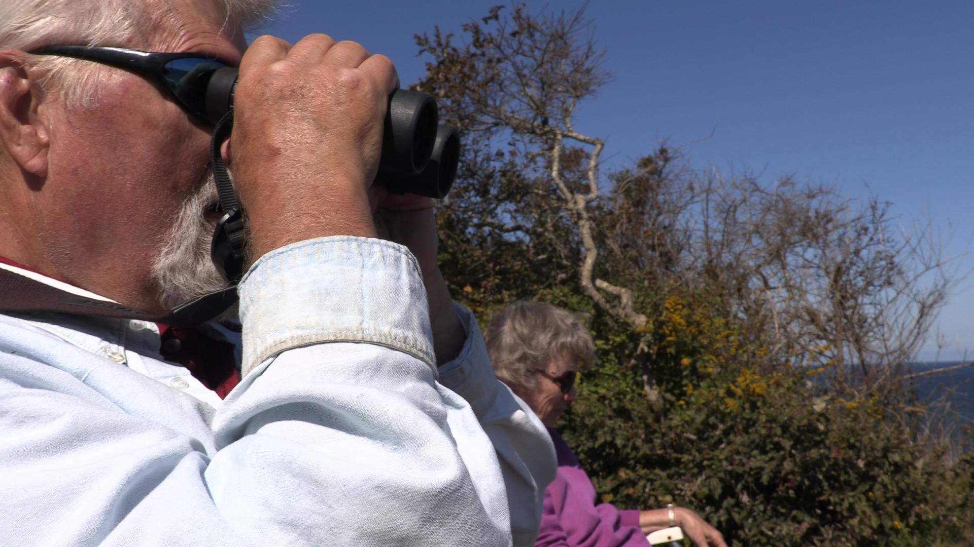 Man sits with his wife and looks through binoculars at the sea.