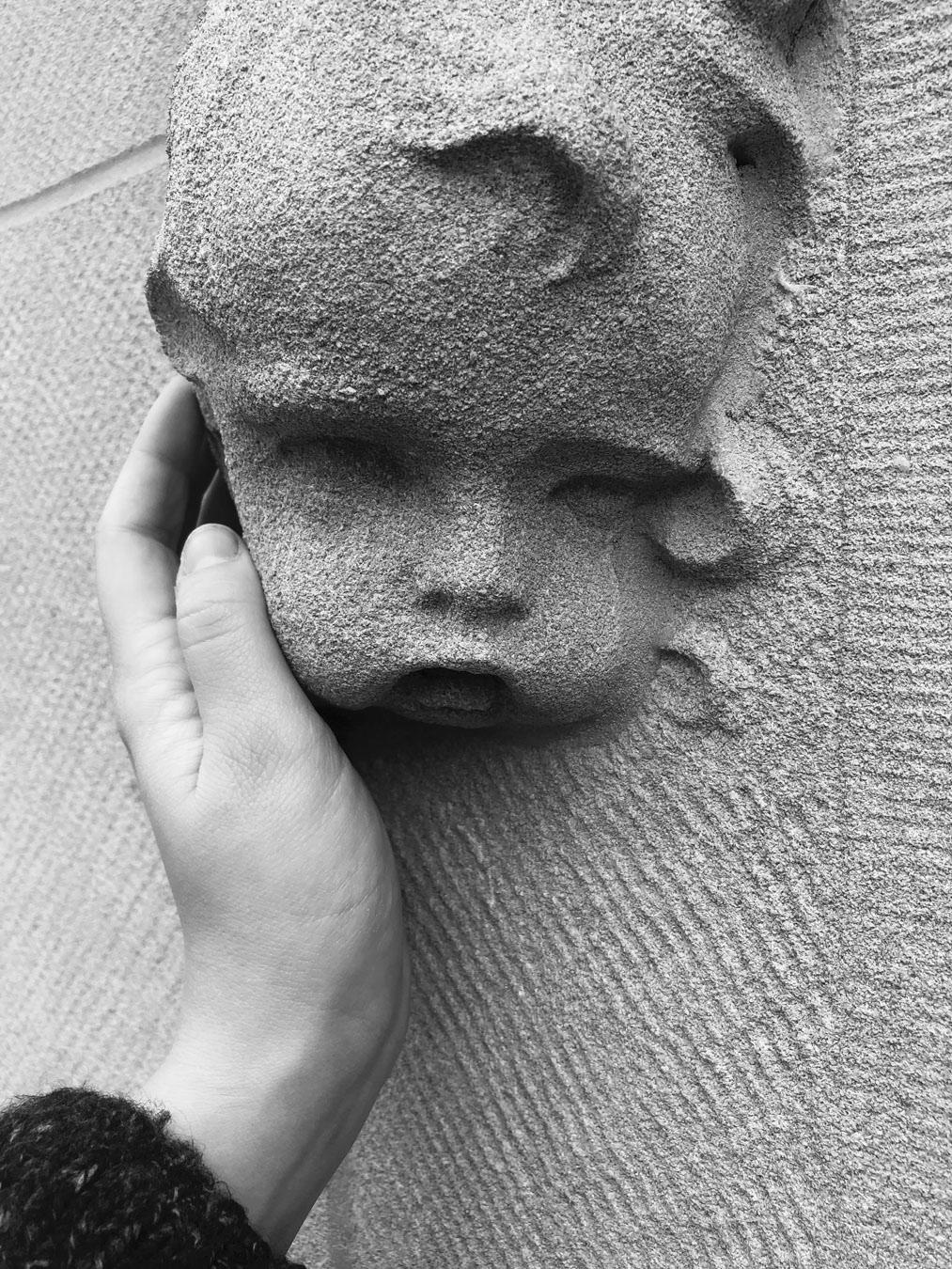 A black and white photograph of a white person's hand reaching out to touch a cherubic face made of stone.