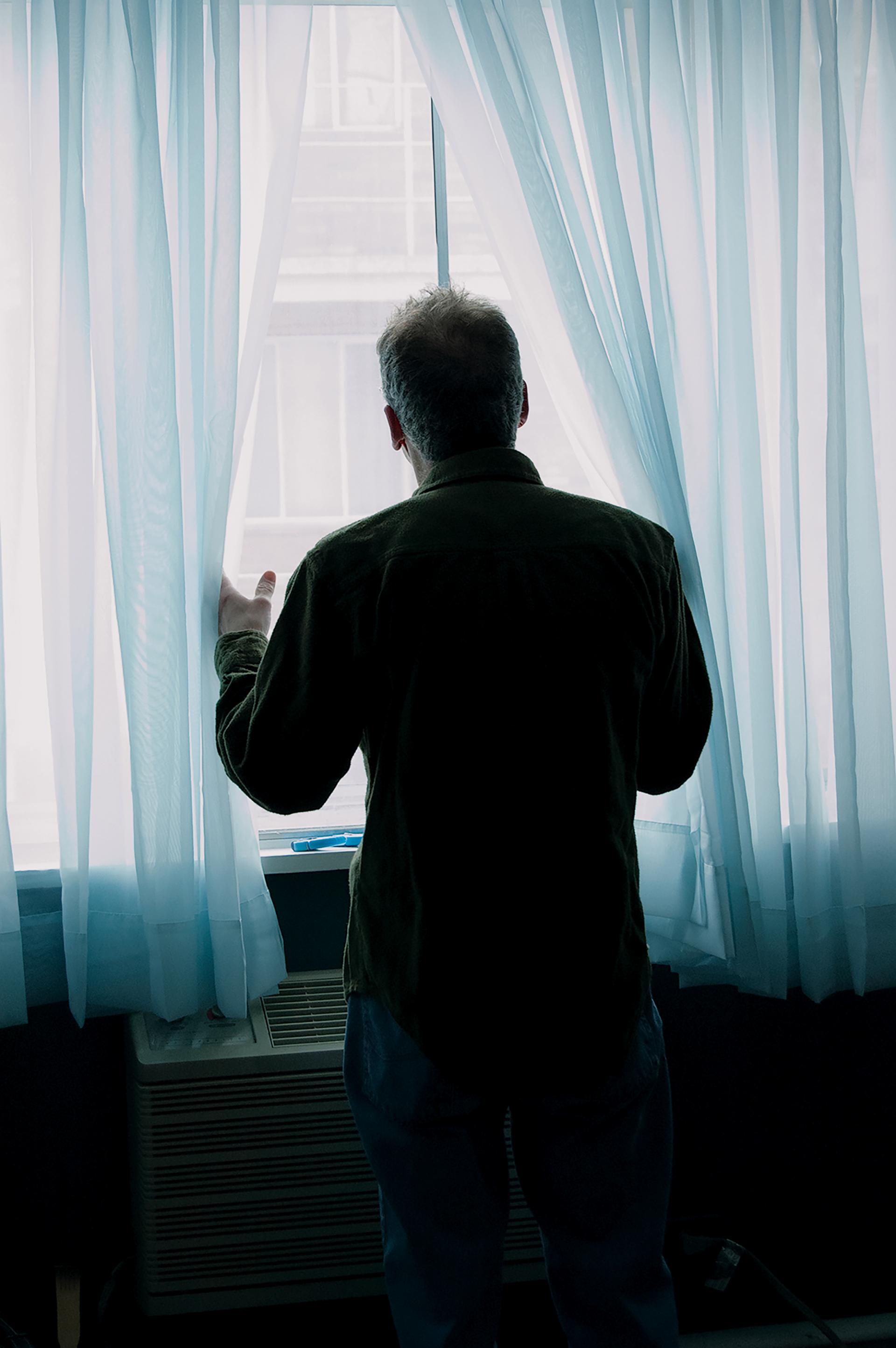 A color photograph of a person looking through translucent blue curtains, seen from behind. 