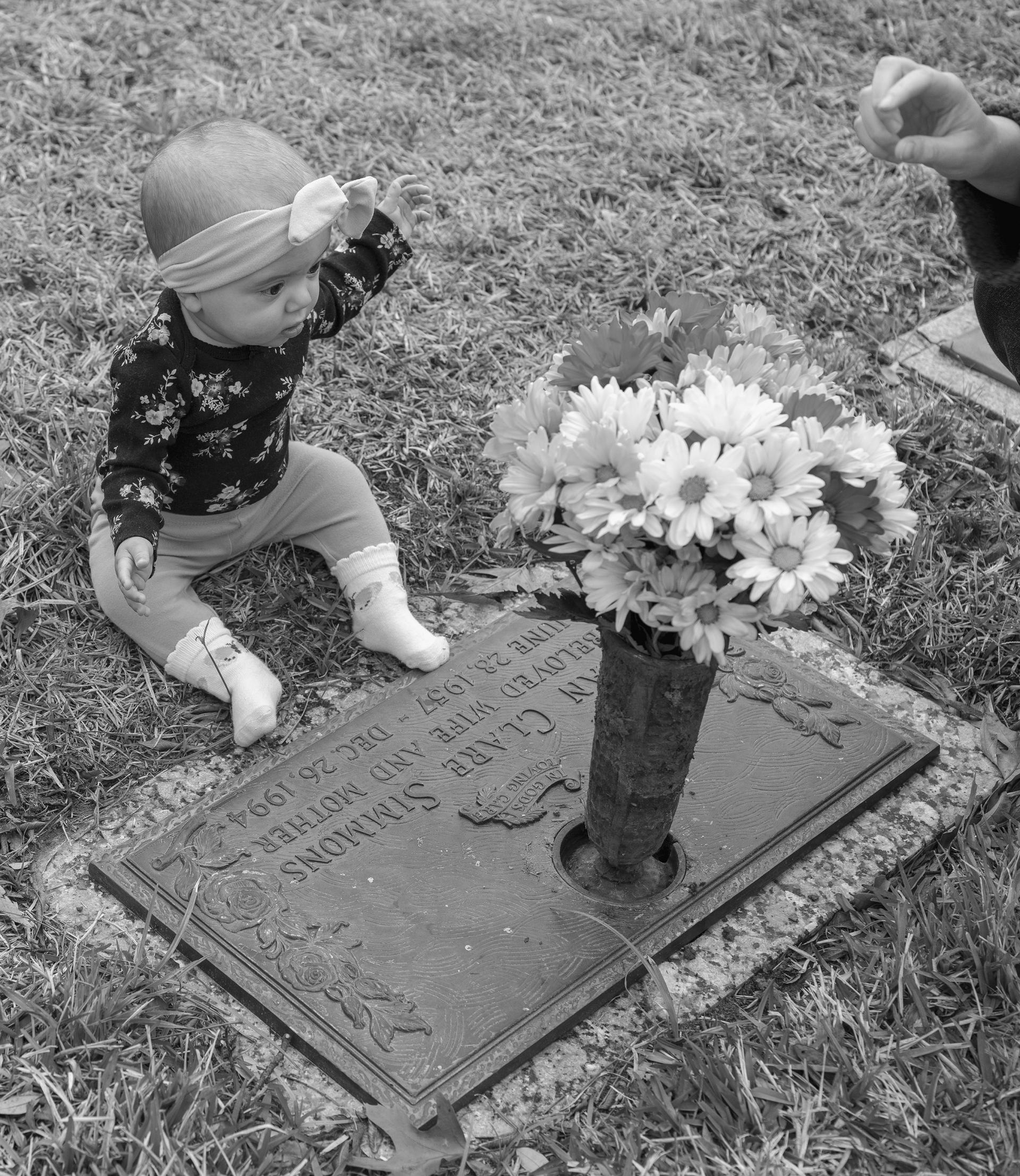 A black and white photograph of a white 6-month-old baby wearing a floral top and a bow, sitting behind a metal headstone. The headstone has a vase on top with multicolored gerbera daisies in it. A white hand is coming into the frame on the right hand side, only visible up to the wrist.