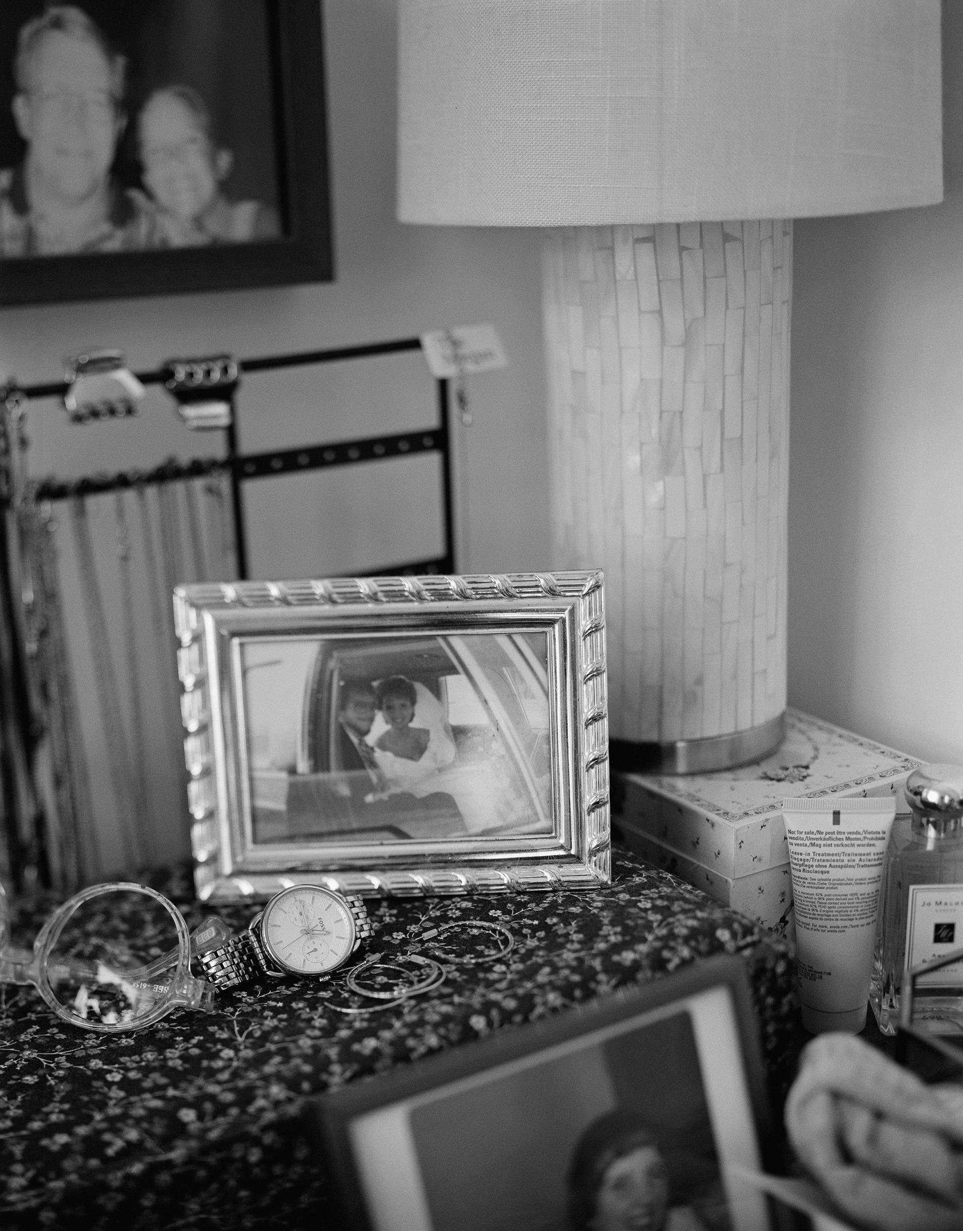 Black and white photograph of a wedding picture from the 1980s in a silver frame, on top of a dresser. 