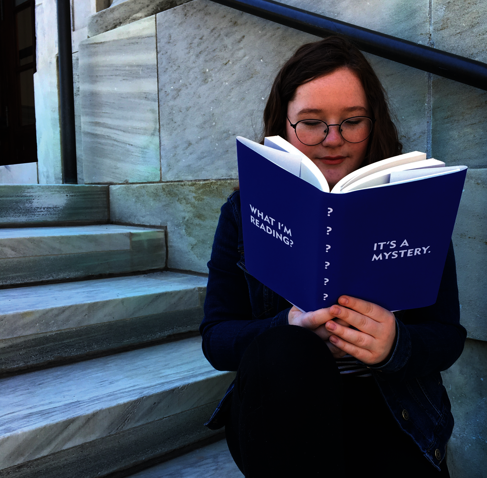 Woman reading a book with a blue cover with text 'What I'm Reading? It's a Mystery'