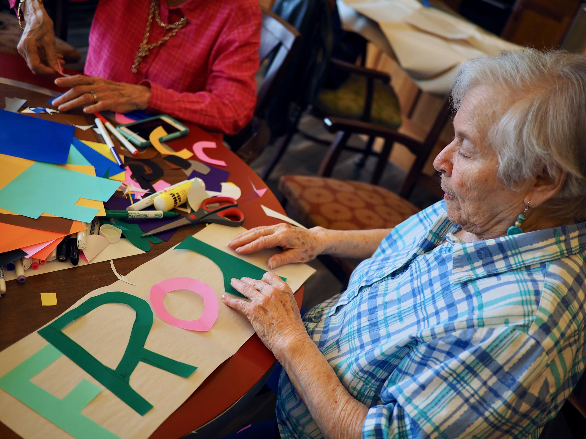 Image of an older female participant, sitting at a table and arranging brightly colored, cutout letters on a poster.