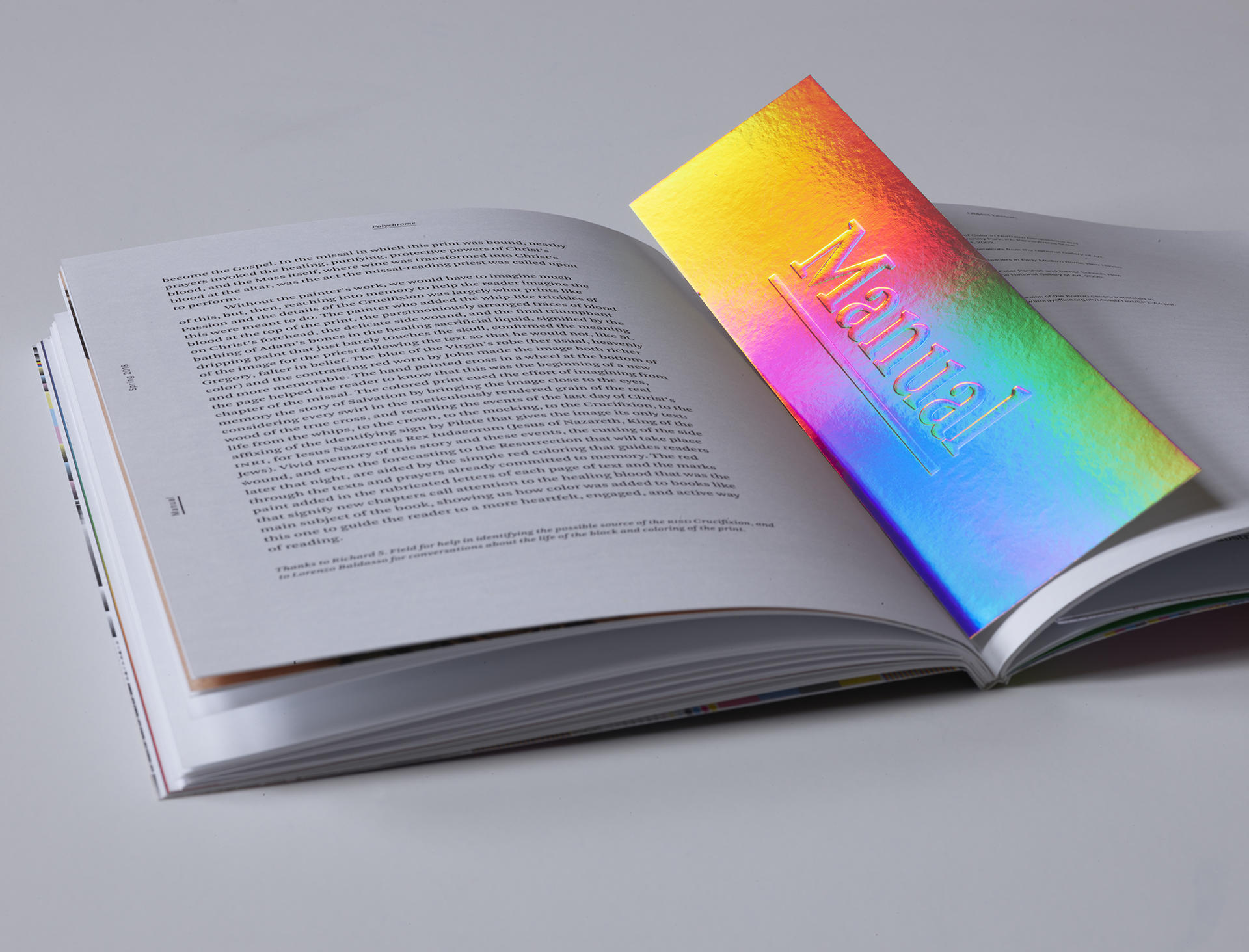 A publication lies open on a white surface. Resting in the middle of the page is a rainbow-colored bookmark, the word “Manual” stamped onto it.