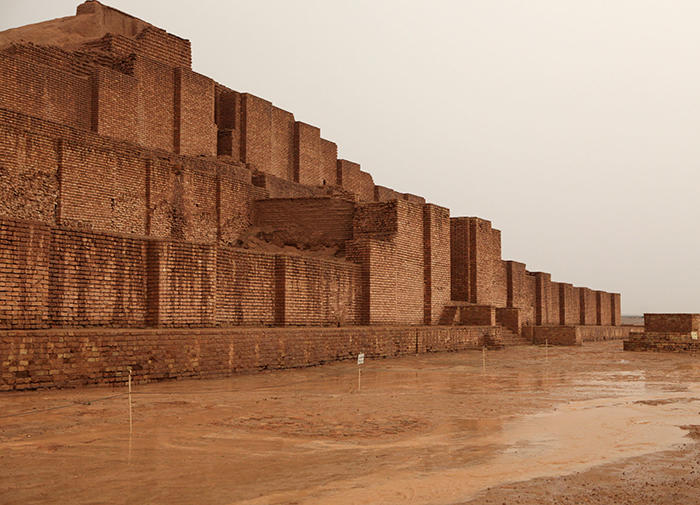 color photograph of the Choqa Zanbil ziggurat. The stepped, square building made of beige stone rises up imposingly from the beige sand.